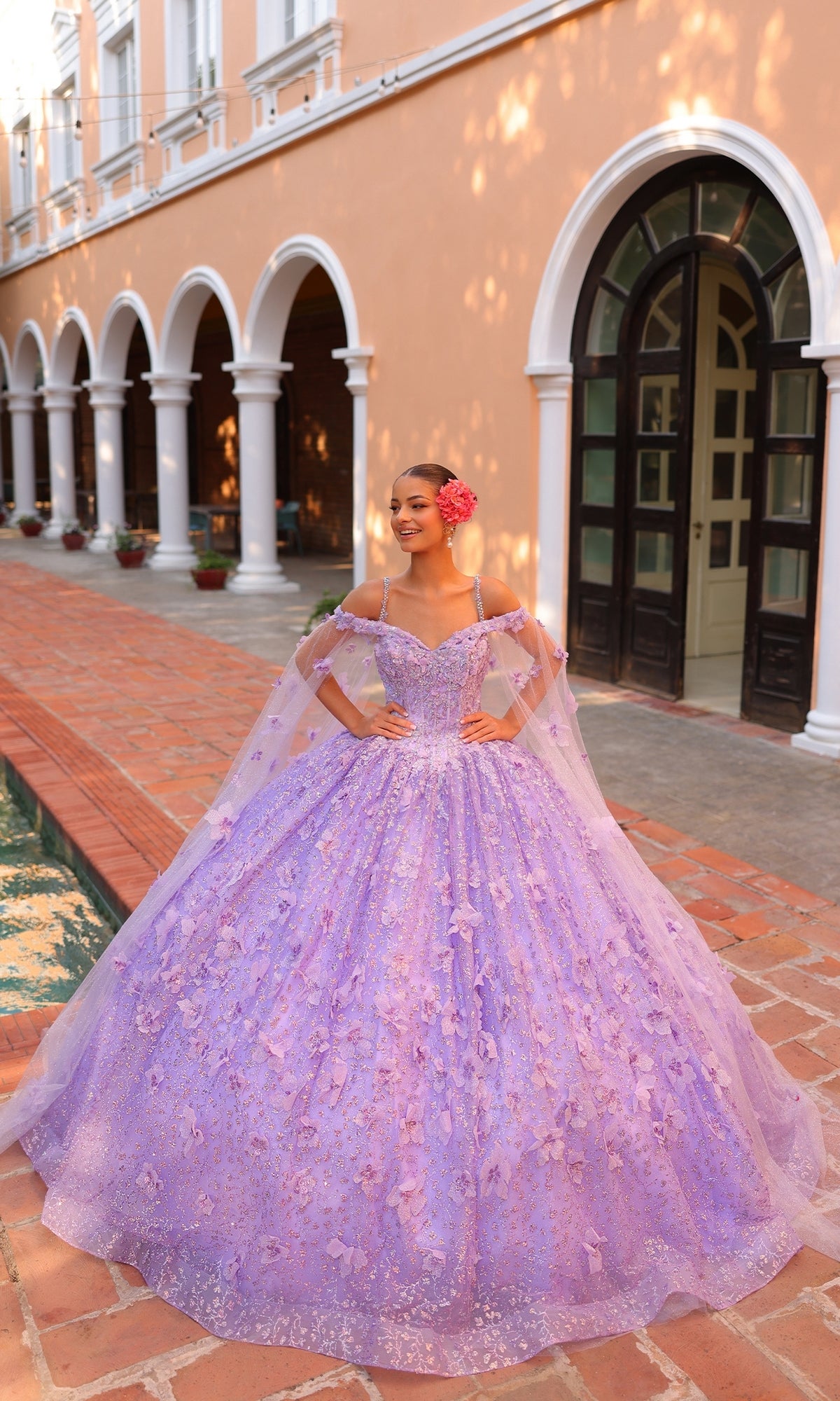 A young woman smiles and poses with her hands on her waist, wearing the Quinceanera Dress 54308 By Amarra?an off-the-shoulder ball gown with 3D floral details?while standing on a brick path in front of an arched building.
