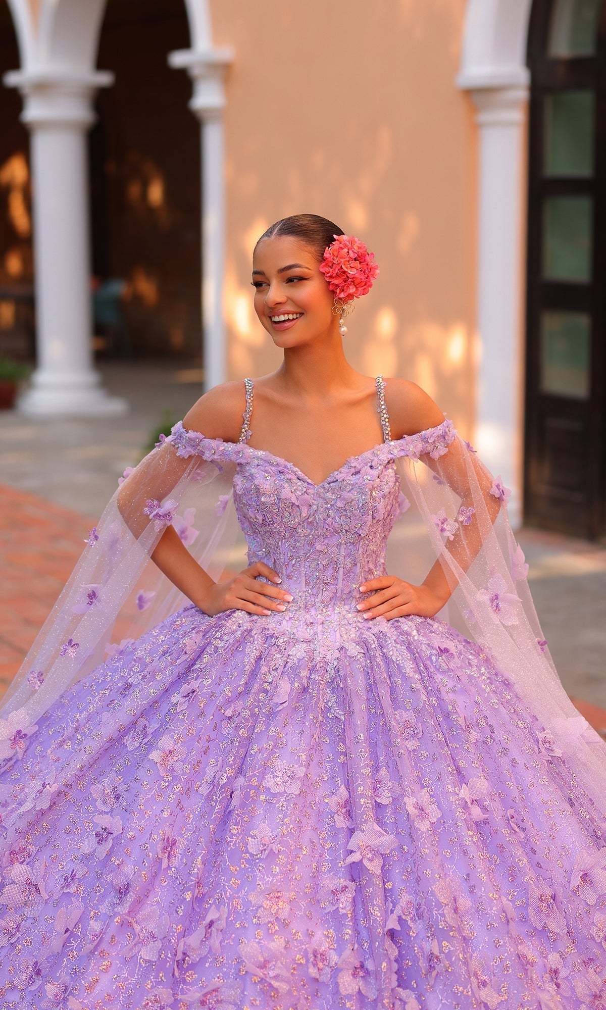 A young woman smiles outside in the Quinceanera Dress 54308 By Amarra, an off-the-shoulder ball gown with 3D floral details, a sheer cape, and a large pink flower in her hair.