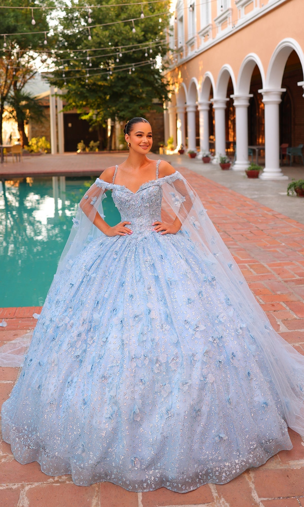A young woman stands by a pool in Quinceanera Dress 54308 By Amarra, featuring off-the-shoulder design, 3D florals, and sheer cape sleeves, set in a courtyard with arches and glowing string lights.