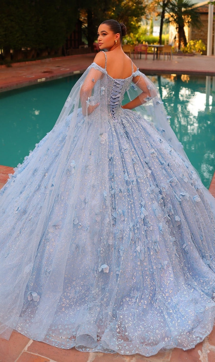 By the pool, a woman wears the Quinceanera Dress 54308 By Amarra?an elegant, sparkly blue off-the-shoulder ball gown featuring 3D floral details and a lace-up back.