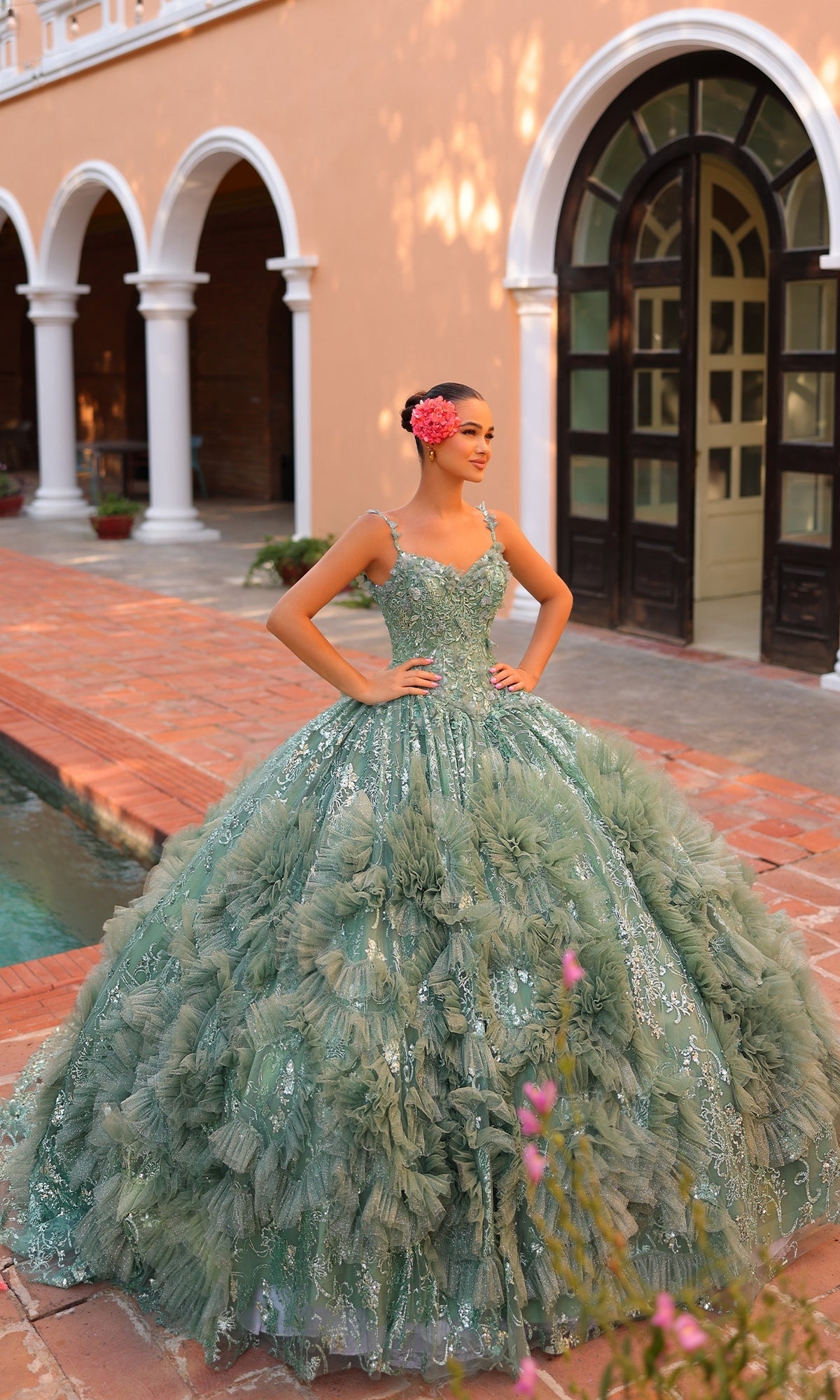 A young woman stands with hands on hips near an arched doorway outdoors, wearing the green, ruffled Quinceanera Dress 54314 by Amarra on a brick patio.