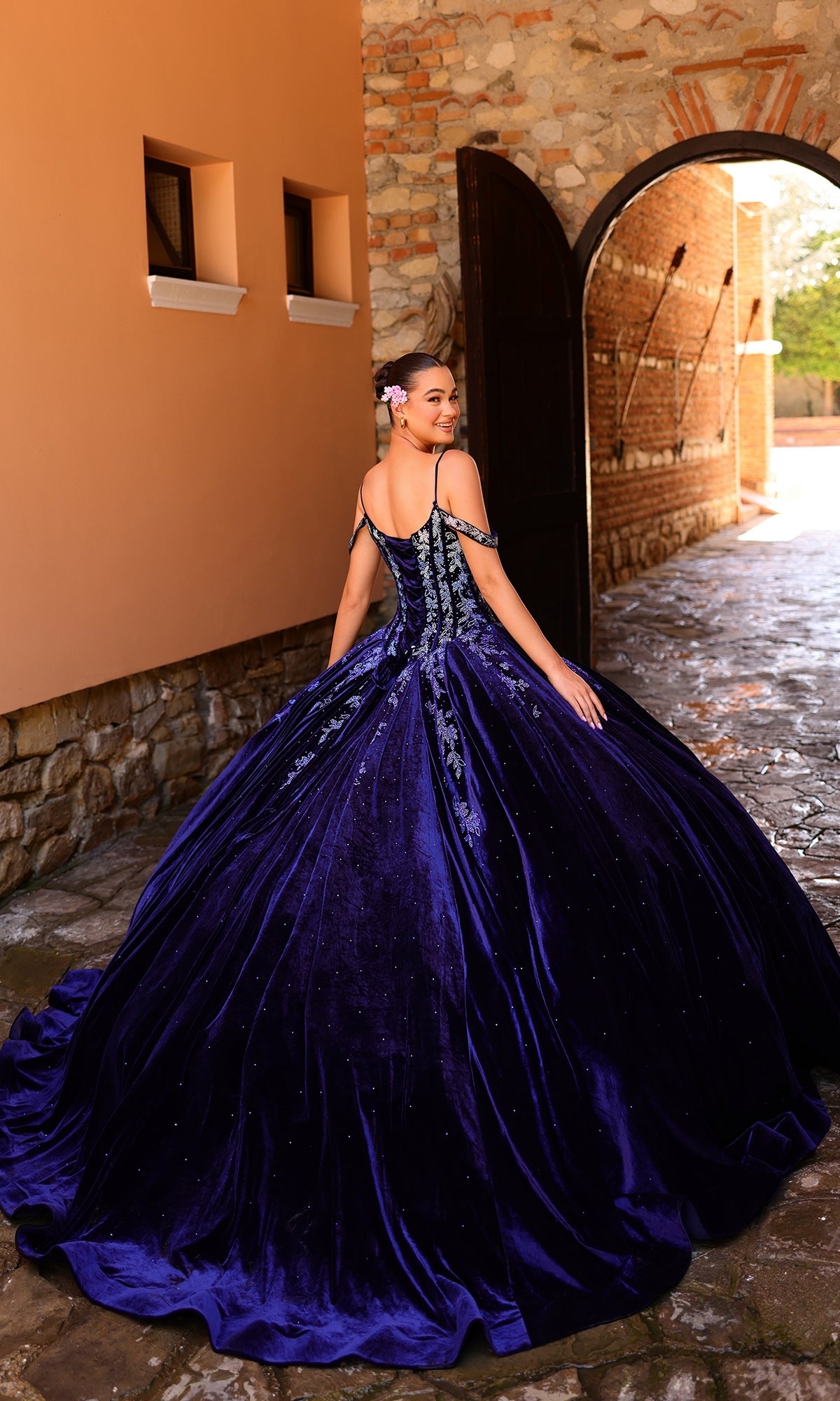 A woman stands outdoors before an arched doorway, wearing the Quinceanera Dress 54316 by Amarra with off-the-shoulder straps and floral hair accessories, looking back over her shoulder.