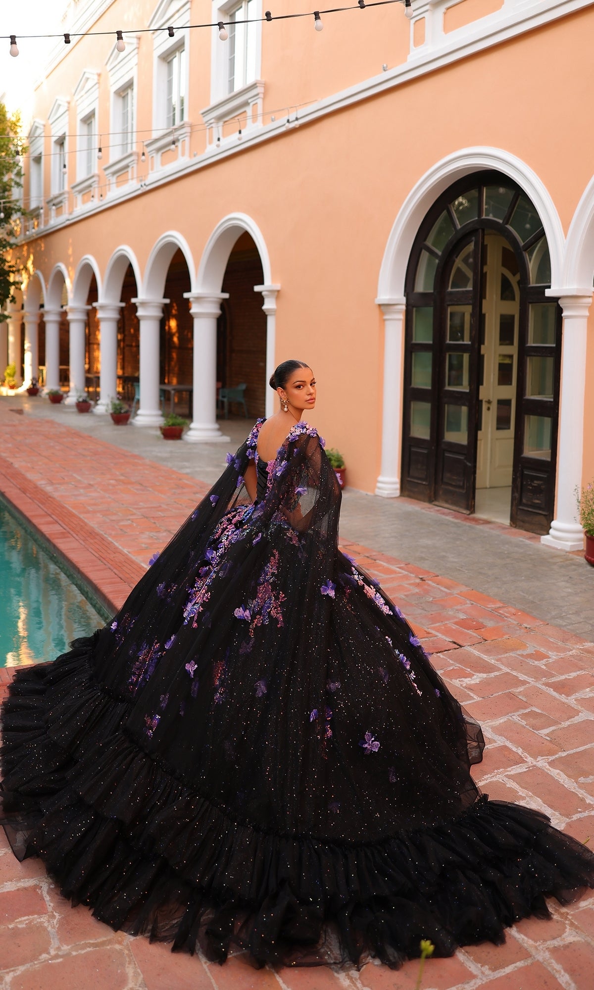 A woman in the Quinceanera Dress 54318 By Amarra, a voluminous black ball gown with purple floral embroidery, stands by a reflecting pool outside an arched building with peach-colored walls.