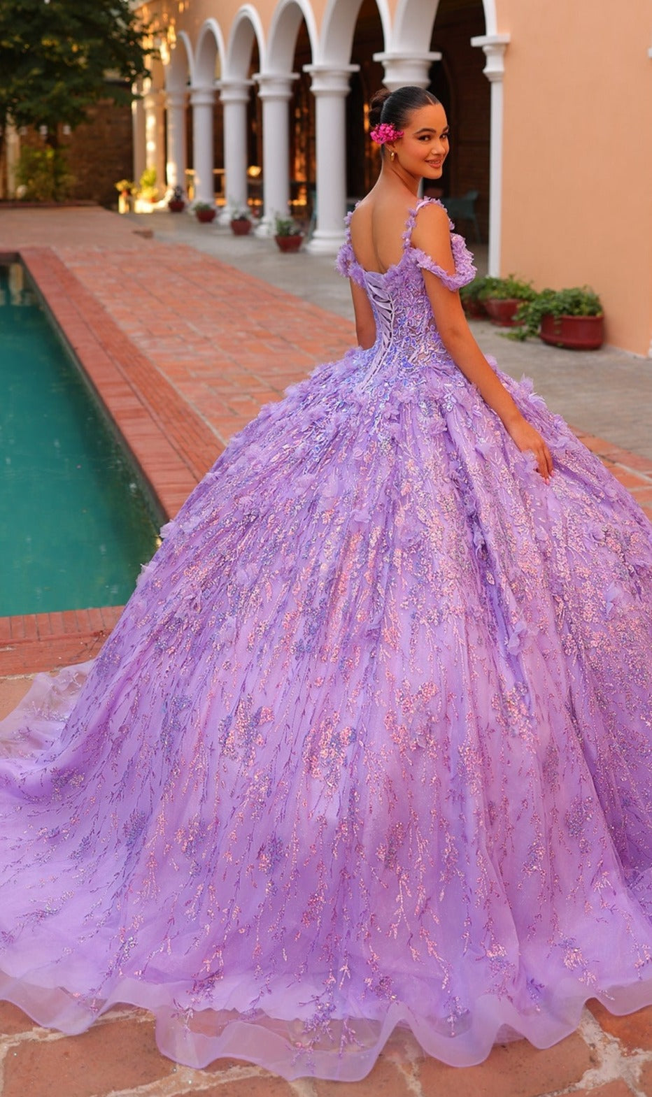 A young woman in the Quinceanera Dress 54320 By Amarra, featuring off-the-shoulder styling and 3D floral details, stands by a pool, smiling over her shoulder.