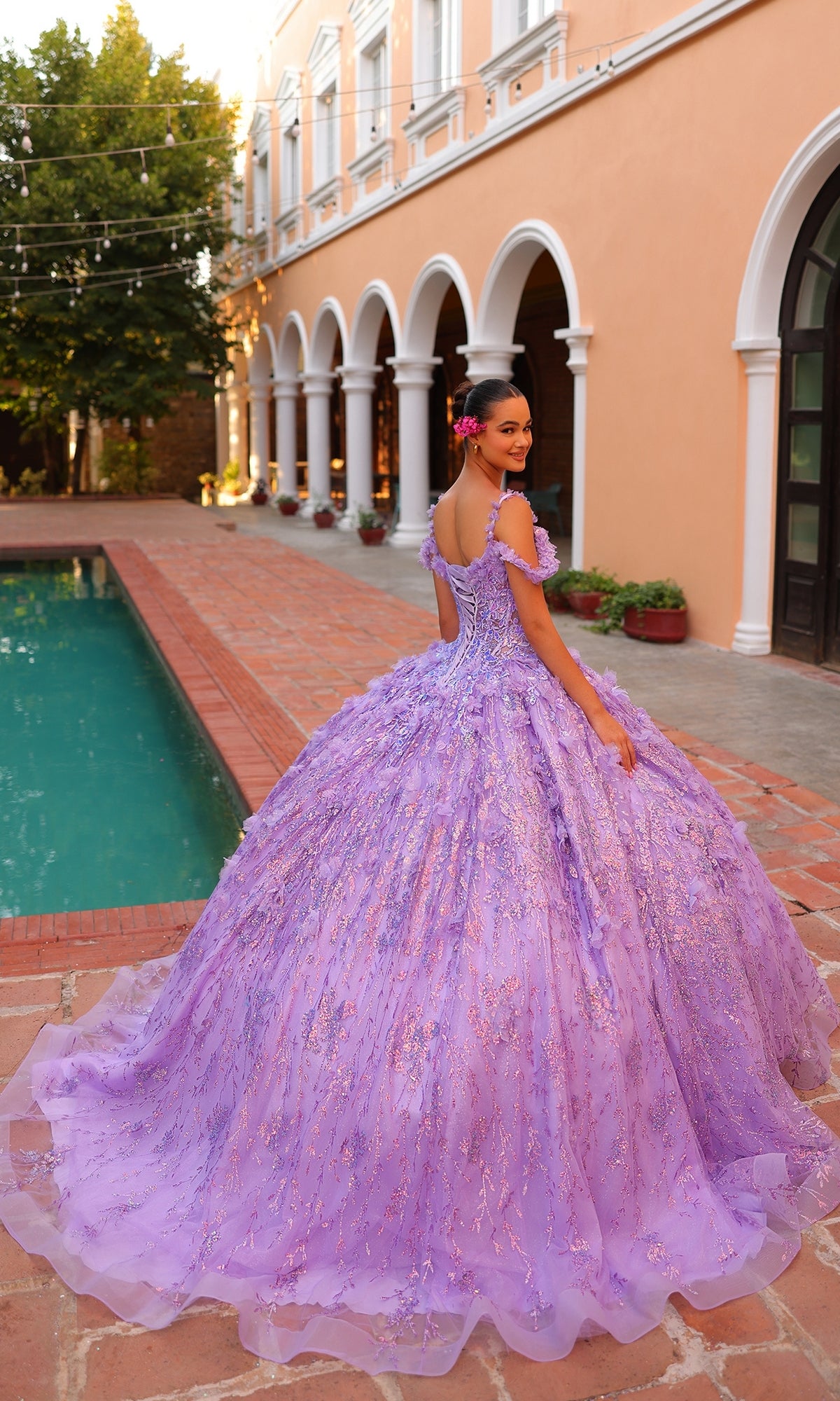 A young woman in the Quinceanera Dress 54320 By Amarra, an off-the-shoulder lavender ball gown, stands by a pool in a courtyard with arched windows, smiling as she looks back over her shoulder.