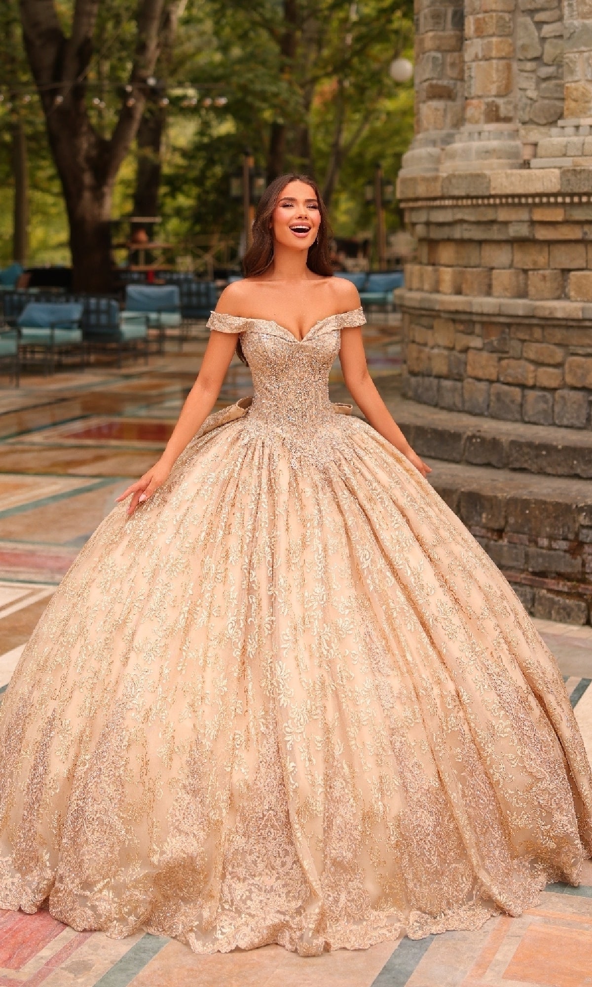 A woman smiles outdoors in the Quinceanera Dress 54324 by Amarra—a gold, off-the-shoulder ball gown with delicate lace embroidery—standing on a patterned stone floor with trees and a stone building behind her.