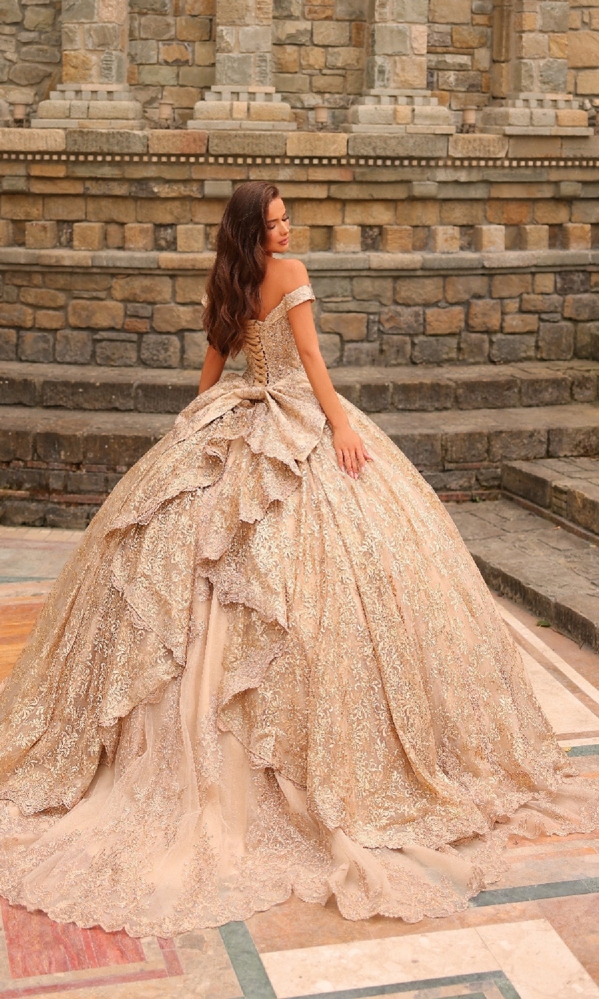 A woman stands outdoors on patterned stone pavement wearing the Quinceanera Dress 54324 By Amarra, a layered, glittering gold ball gown with off-the-shoulder sleeves; a stone wall is in the background.