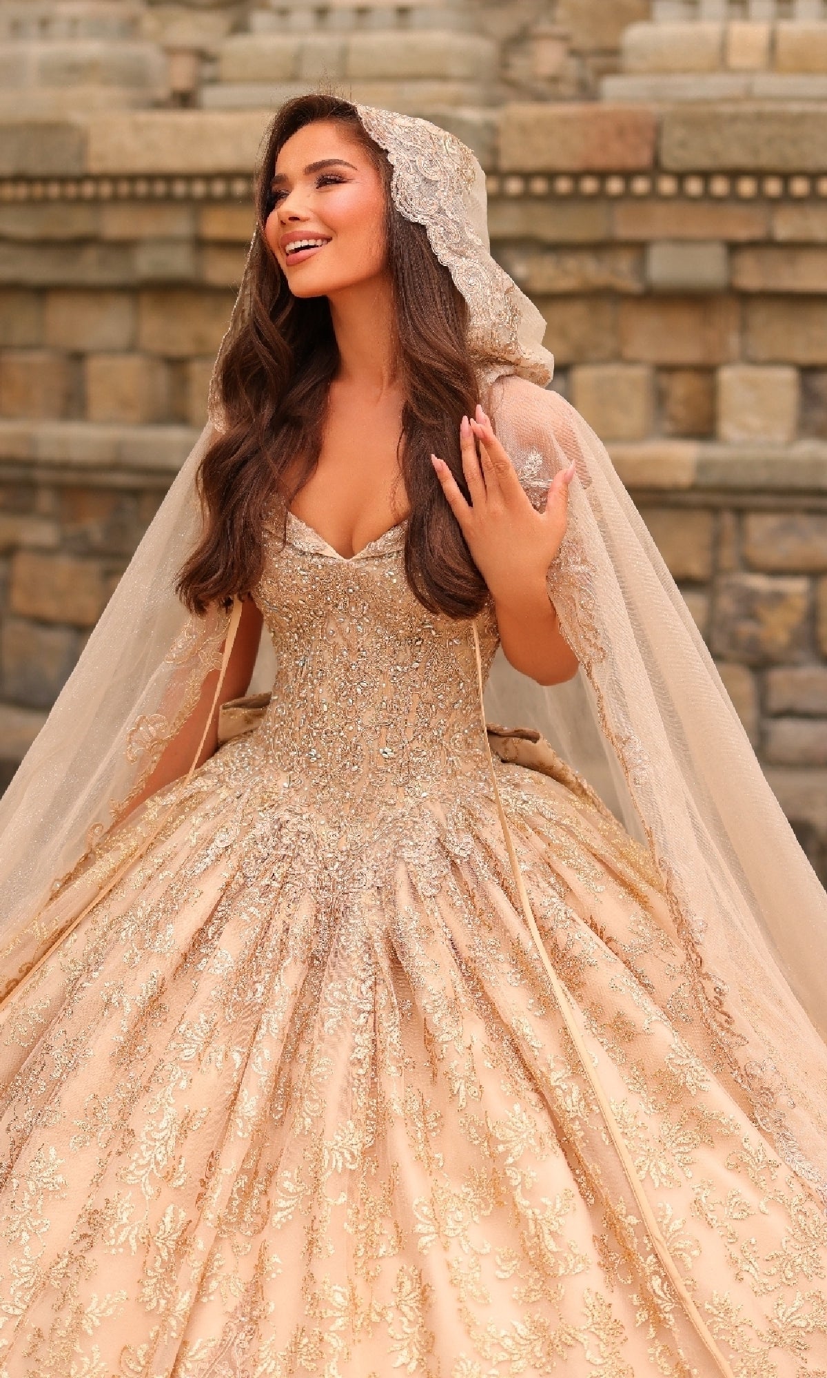 A woman smiles in front of a stone wall, wearing the Quinceanera Dress 54324 By Amarra—an off-the-shoulder gold ball gown with lace embroidery and a matching veil, holding part of her veil.