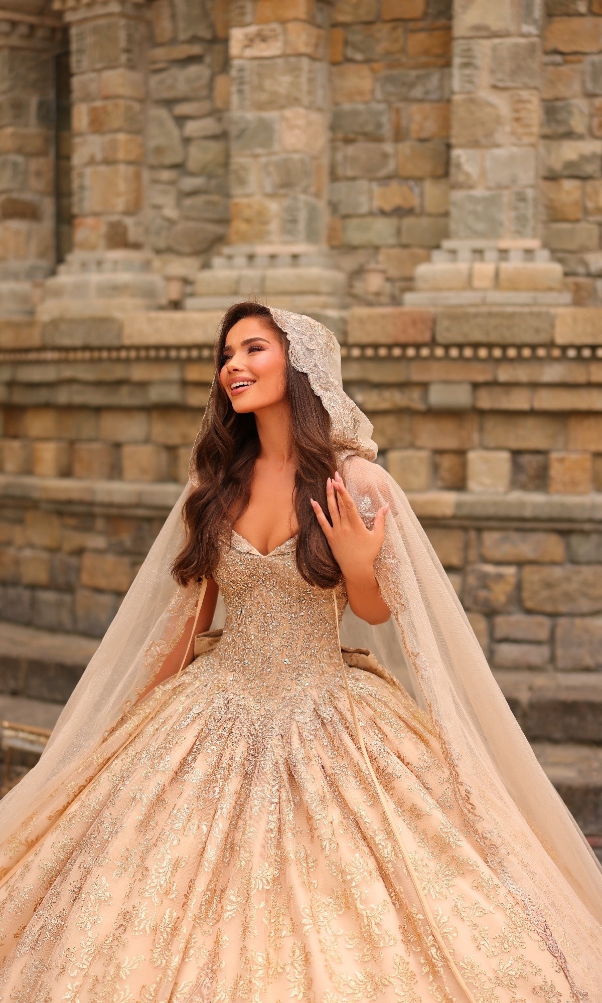 A woman wearing the Quinceanera Dress 54324 by Amarra, featuring gold-embroidered lace and an off-the-shoulder design with a hooded cape, poses smiling in front of a stone wall.
