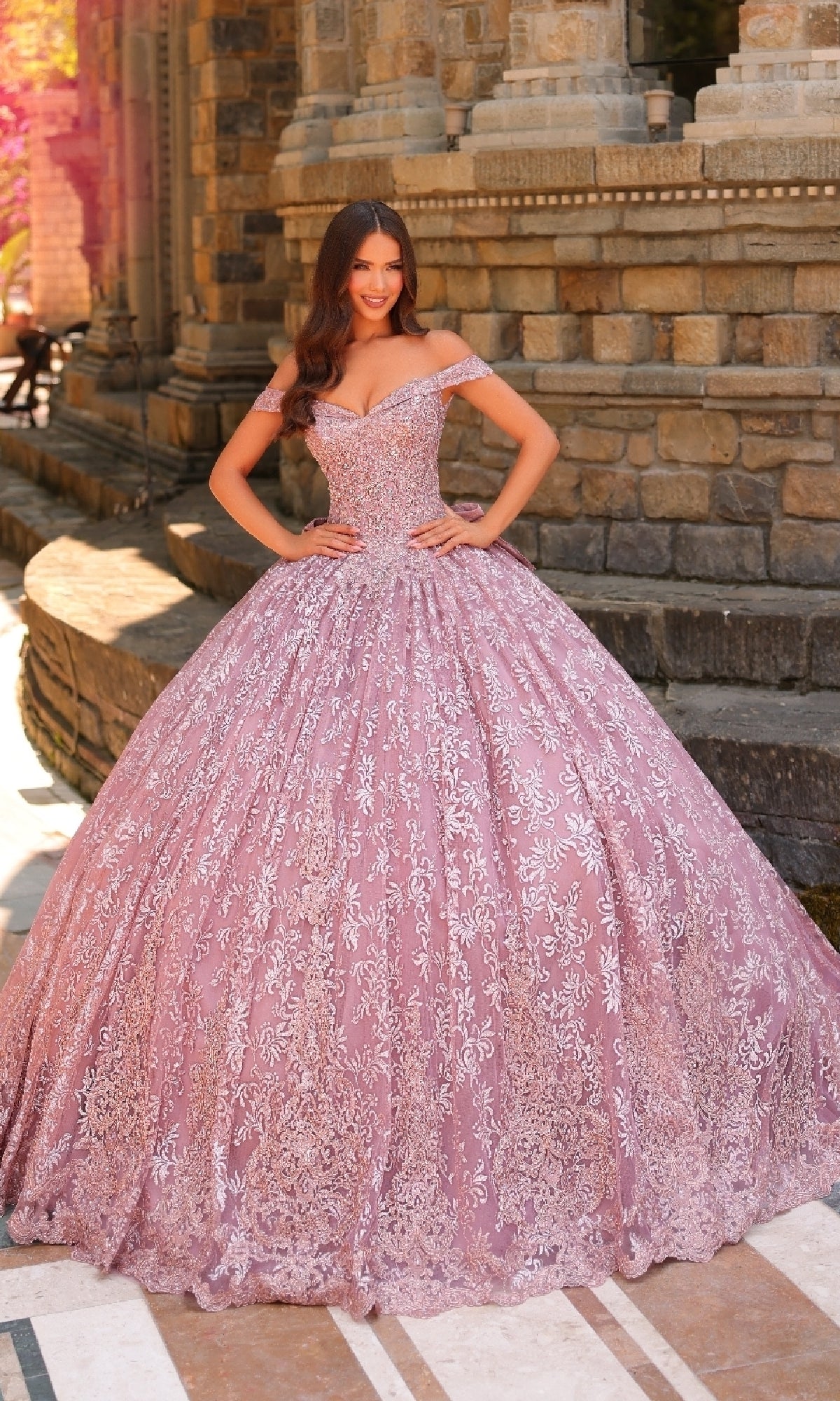 A woman stands outdoors in front of a stone building wearing the Quinceanera Dress 54324 By Amarra, an ornate off-the-shoulder ball gown featuring silver floral patterns and delicate lace embroidery.
