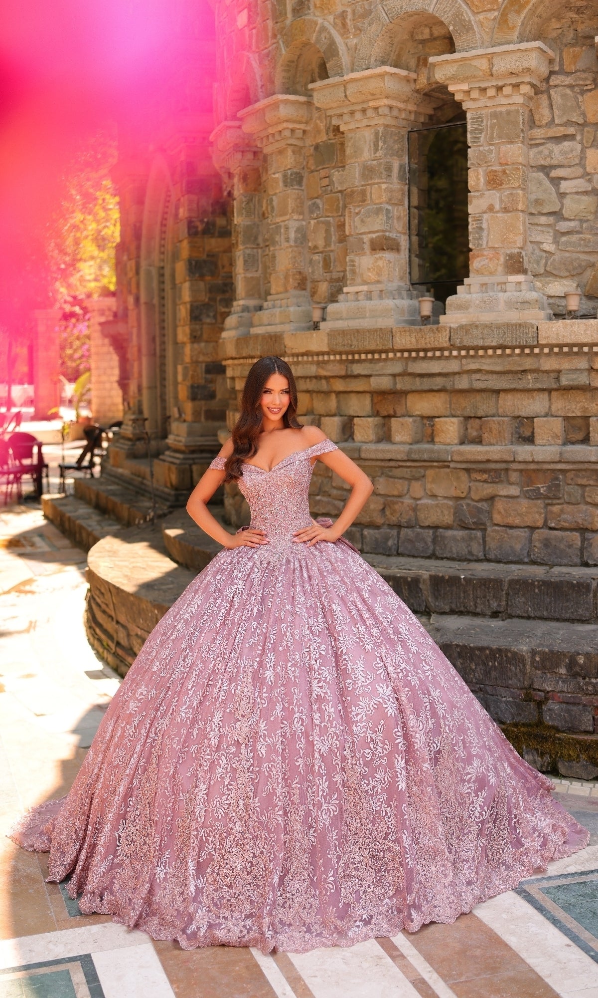 A woman stands outdoors in front of a stone building, wearing the Quinceanera Dress 54324 by Amarra—a pink ball gown featuring off-the-shoulder sleeves, intricate lace embroidery, and a voluminous skirt.