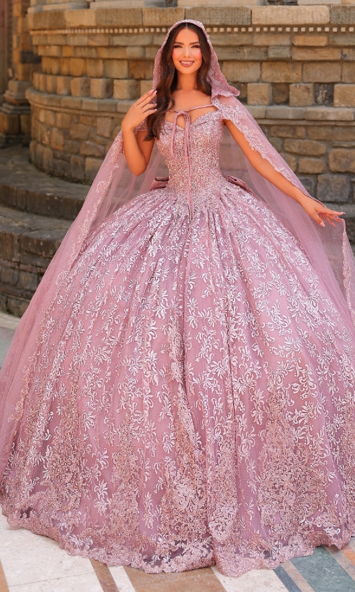 A woman stands before a stone wall in the Quinceanera Dress 54324 by Amarra—a voluminous, light purple ball gown featuring off-the-shoulder sleeves, intricate lace embroidery, and a matching sheer cape.
