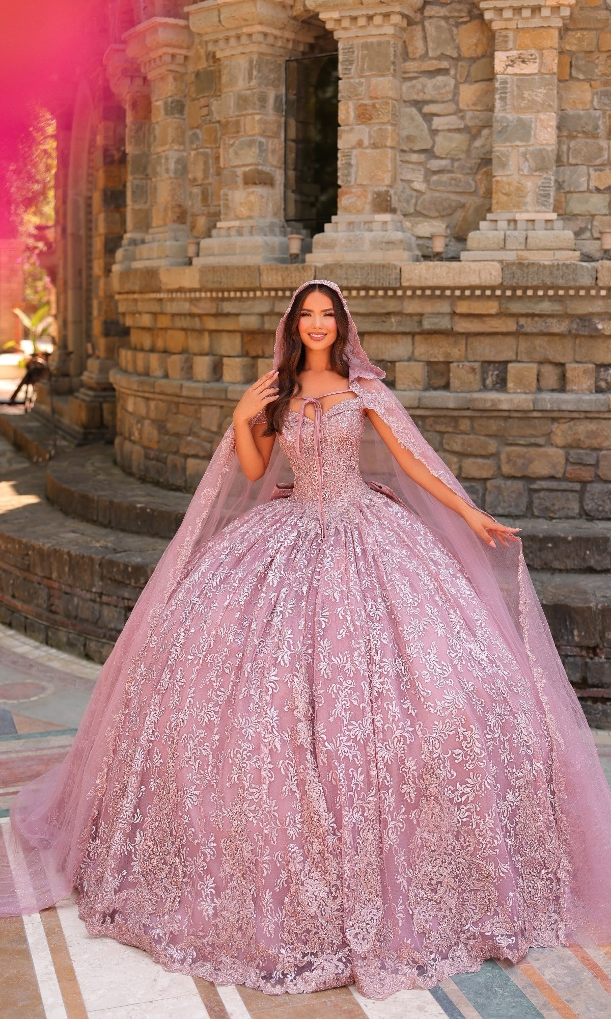 A woman stands outdoors in front of a stone building, wearing the Quinceanera Dress 54324 By Amarra—an ornate, off-the-shoulder light purple ball gown with a matching cape and intricate silver lace embroidery.