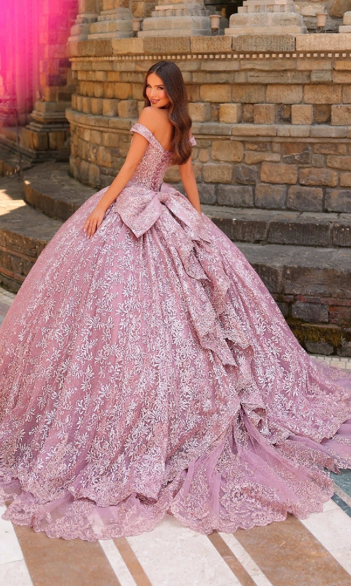 A woman poses outdoors before a stone wall in the Quinceanera Dress 54324 by Amarra, an elaborate off-the-shoulder ball gown featuring intricate lace embroidery and a large back bow.