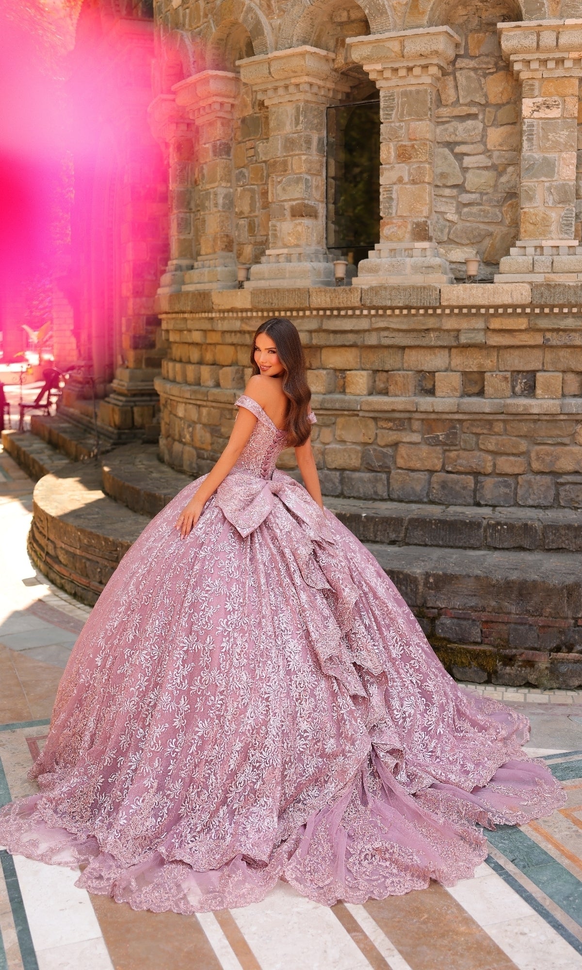 Wearing the Quinceanera Dress 54324 By Amarra with lace embroidery and off-the-shoulder sleeves, a woman stands before a stone building, glancing over her shoulder.