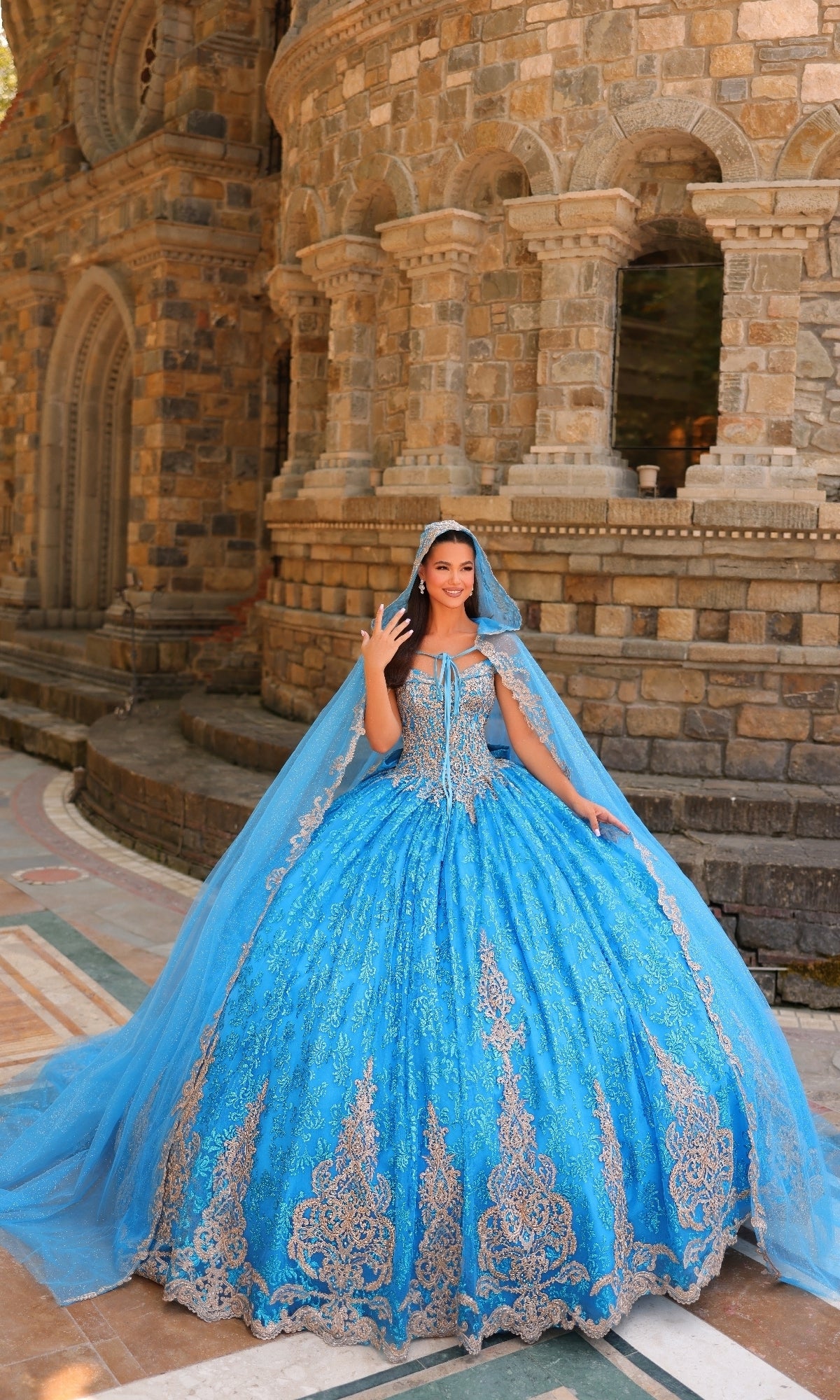 A woman in the Quinceanera Dress 54324 By Amarra—a bright blue, off-the-shoulder ball gown with gold detailing, lace embroidery, and a matching cape—stands gracefully before a stone building with arched windows.