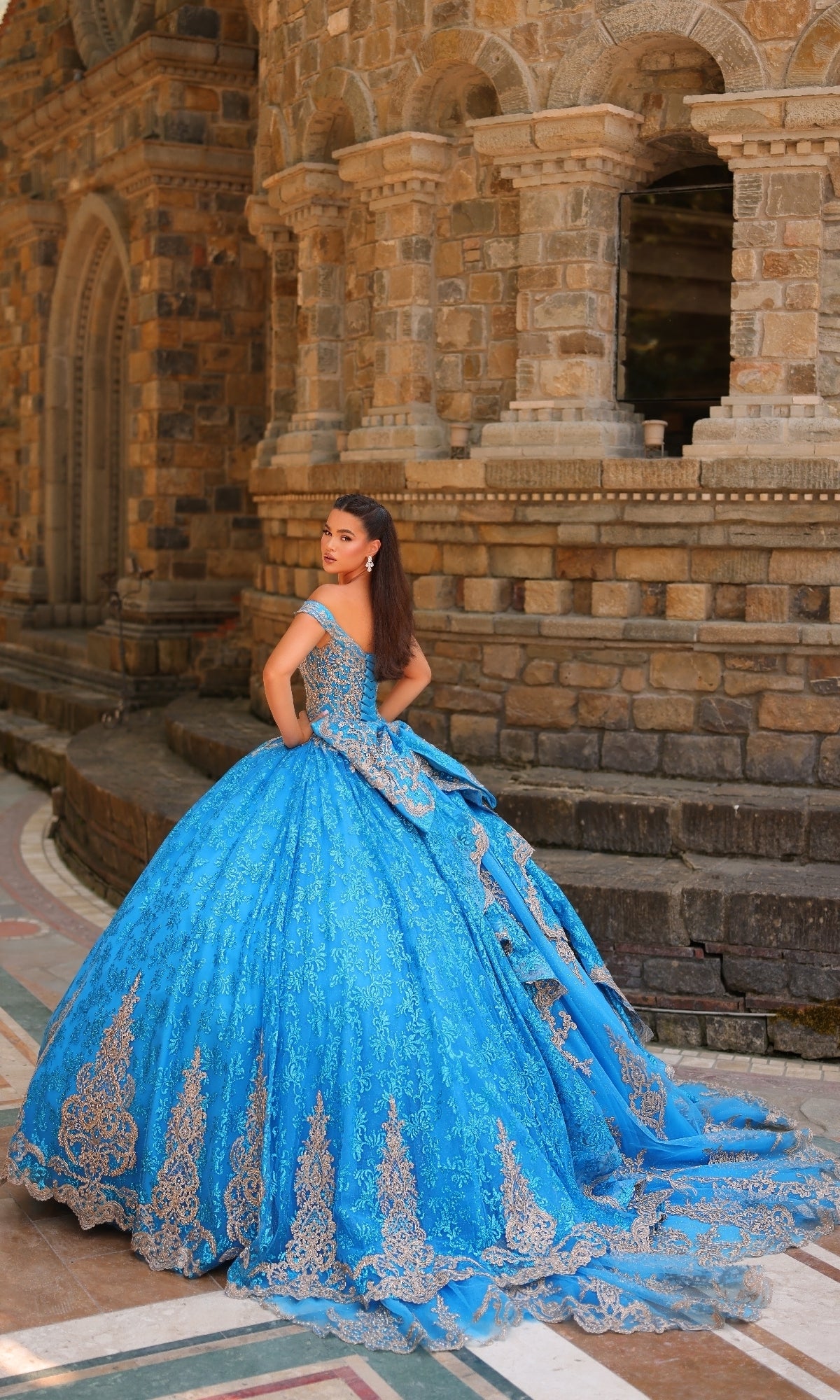 A woman wearing the Quinceanera Dress 54324 by Amarra, a bright blue off-the-shoulder ball gown with silver embellishments, poses in front of a stone building, glancing over her shoulder.