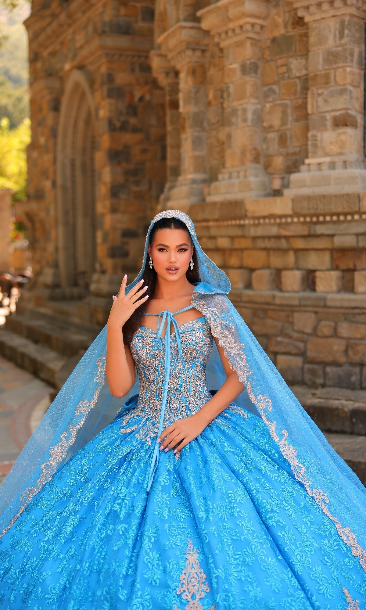 A woman poses in front of a stone building wearing the Quinceanera Dress 54324 by Amarra—an ornate, off-the-shoulder blue ball gown with a matching lace-trimmed cape.