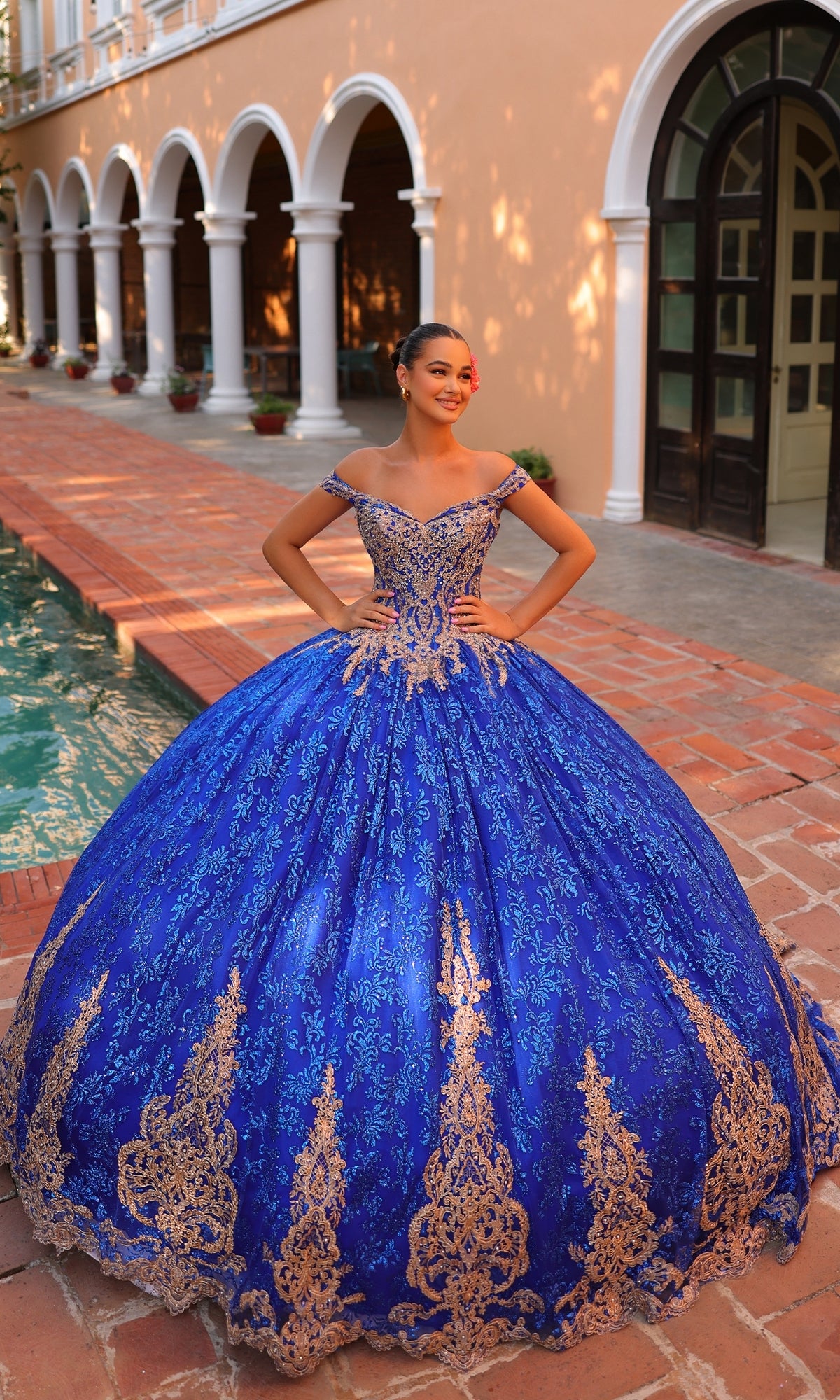 A woman stands by a pool in a courtyard, wearing the Quinceanera Dress 54324 by Amarra, a vibrant blue ball gown featuring gold lace embroidery and off-the-shoulder sleeves.