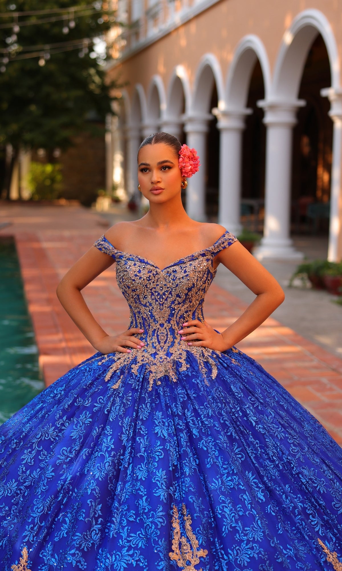 A young woman in the Quinceanera Dress 54324 by Amarra, featuring blue and gold lace embroidery and an off-the-shoulder design, poses by a pool with hands on hips, a pink flower in her hair, and an ornate building behind her.
