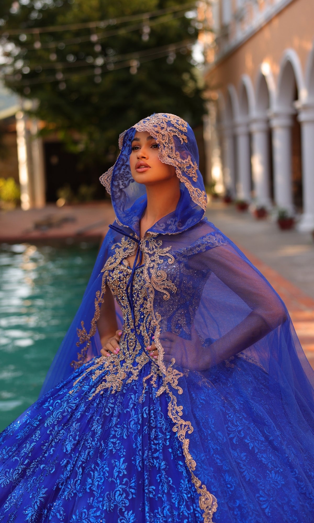 A person stands by a pool in front of an arched building, wearing Quinceanera Dress 54324 By Amarra—an ornate blue and gold ball gown with lace embroidery and a matching hooded cape, under glowing string lights.