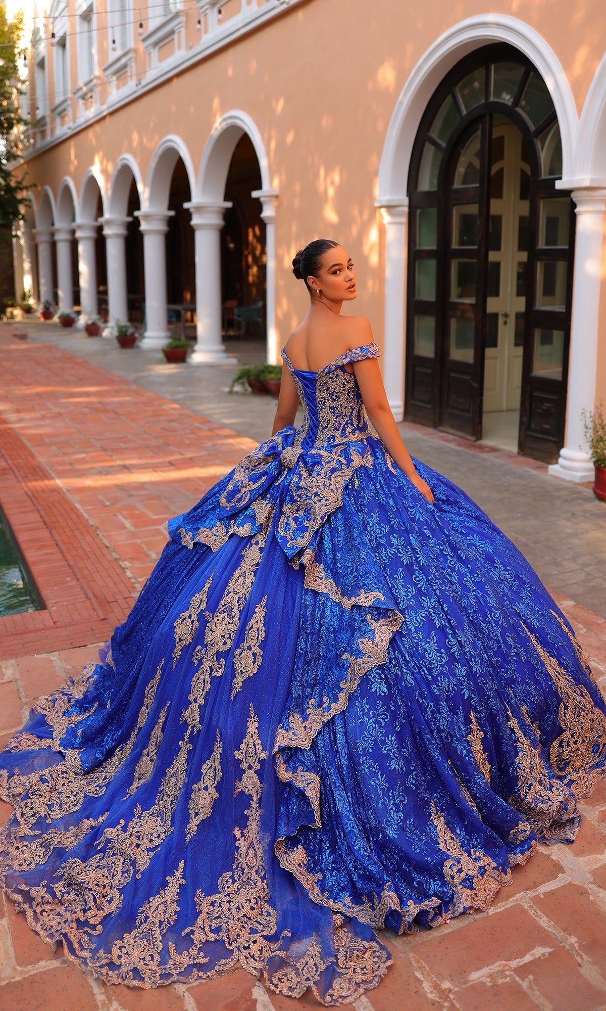 A woman stands in a courtyard wearing the Quinceanera Dress 54324 by Amarra, an elaborate royal blue ball gown with gold lace embroidery, looking over her shoulder.