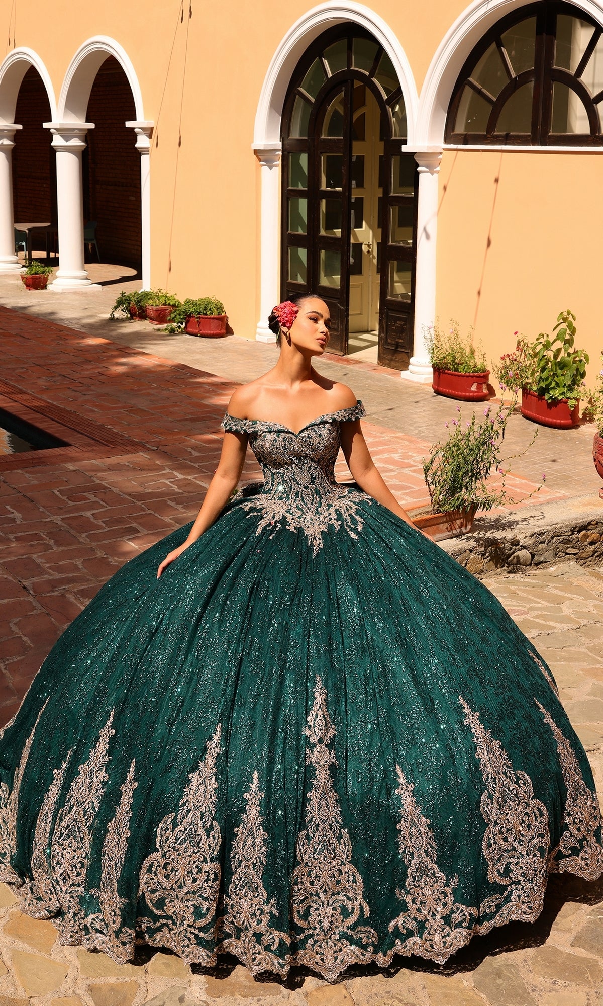 A woman stands outdoors near an arched building with potted plants, showcasing the Quinceanera Dress 54324 By Amarra—a large, ornate off-the-shoulder green ball gown featuring silver lace embroidery.