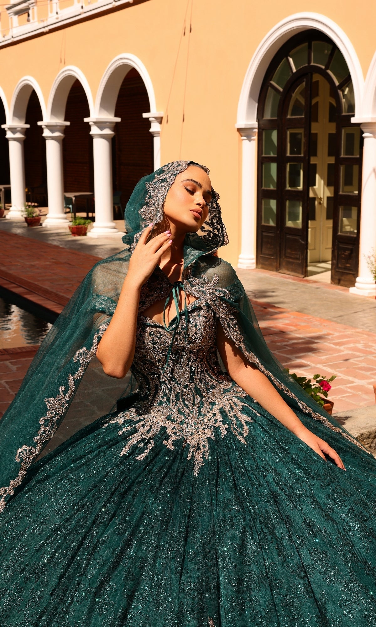 A woman wears the Quinceanera Dress 54324 By Amarra, an off-the-shoulder green gown with silver lace embroidery and a matching hooded cape, posing outdoors before arched doorways.