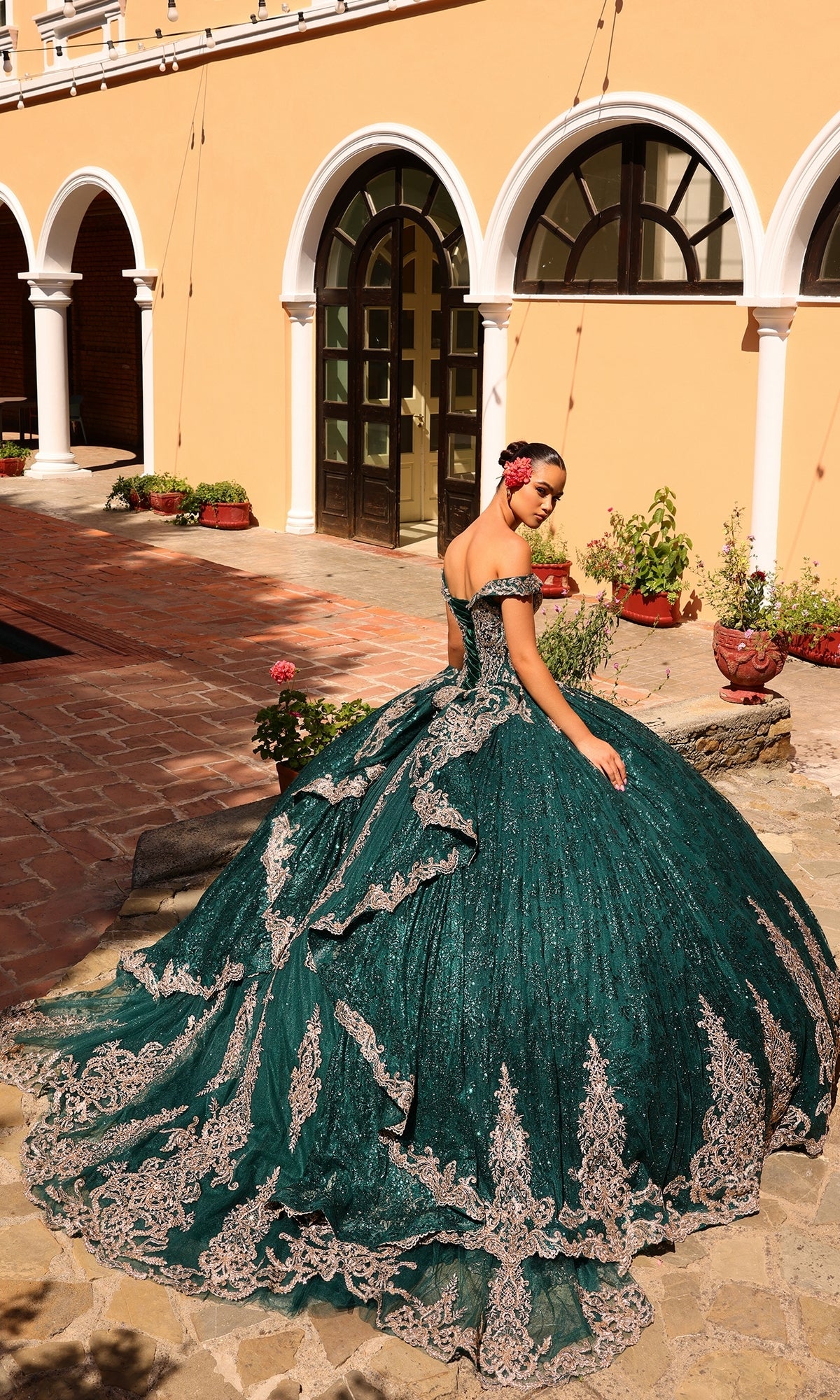 A woman stands outdoors in front of an arched doorway and yellow building, wearing the Quinceanera Dress 54324 by Amarra—an elaborate green ball gown with silver detailing.