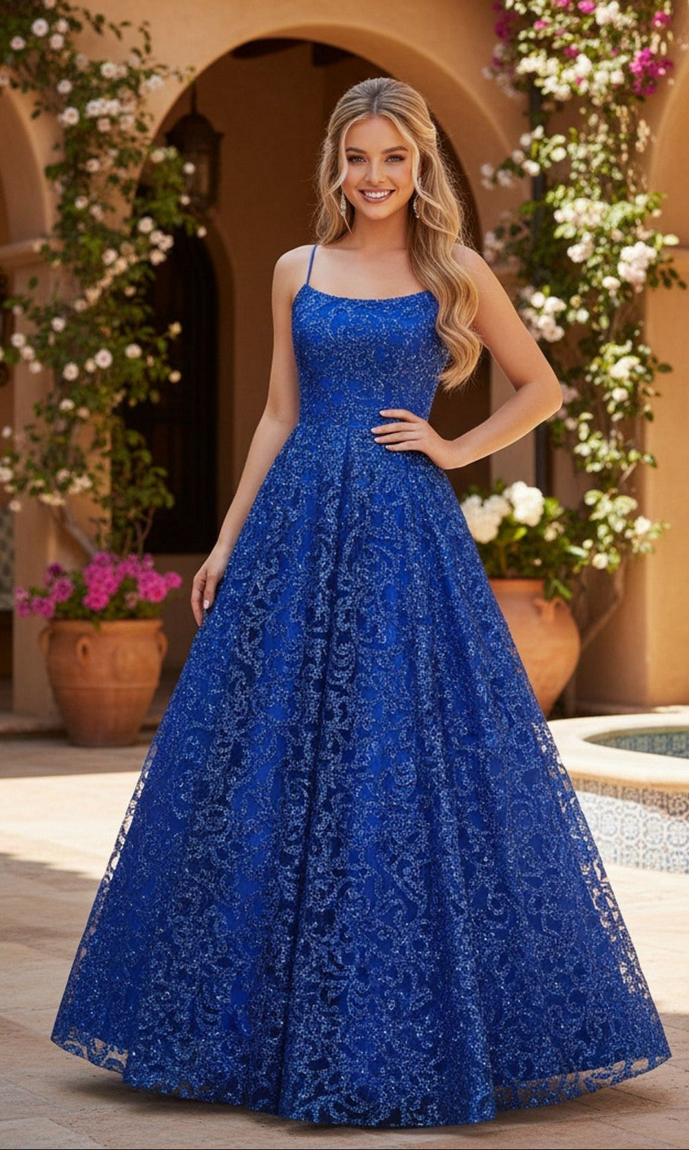 A woman stands outdoors by an arched building and flowering plants, wearing the Blondie Nites 5450BN long prom dress in royal—a floor-length, sleeveless gown with glitter.