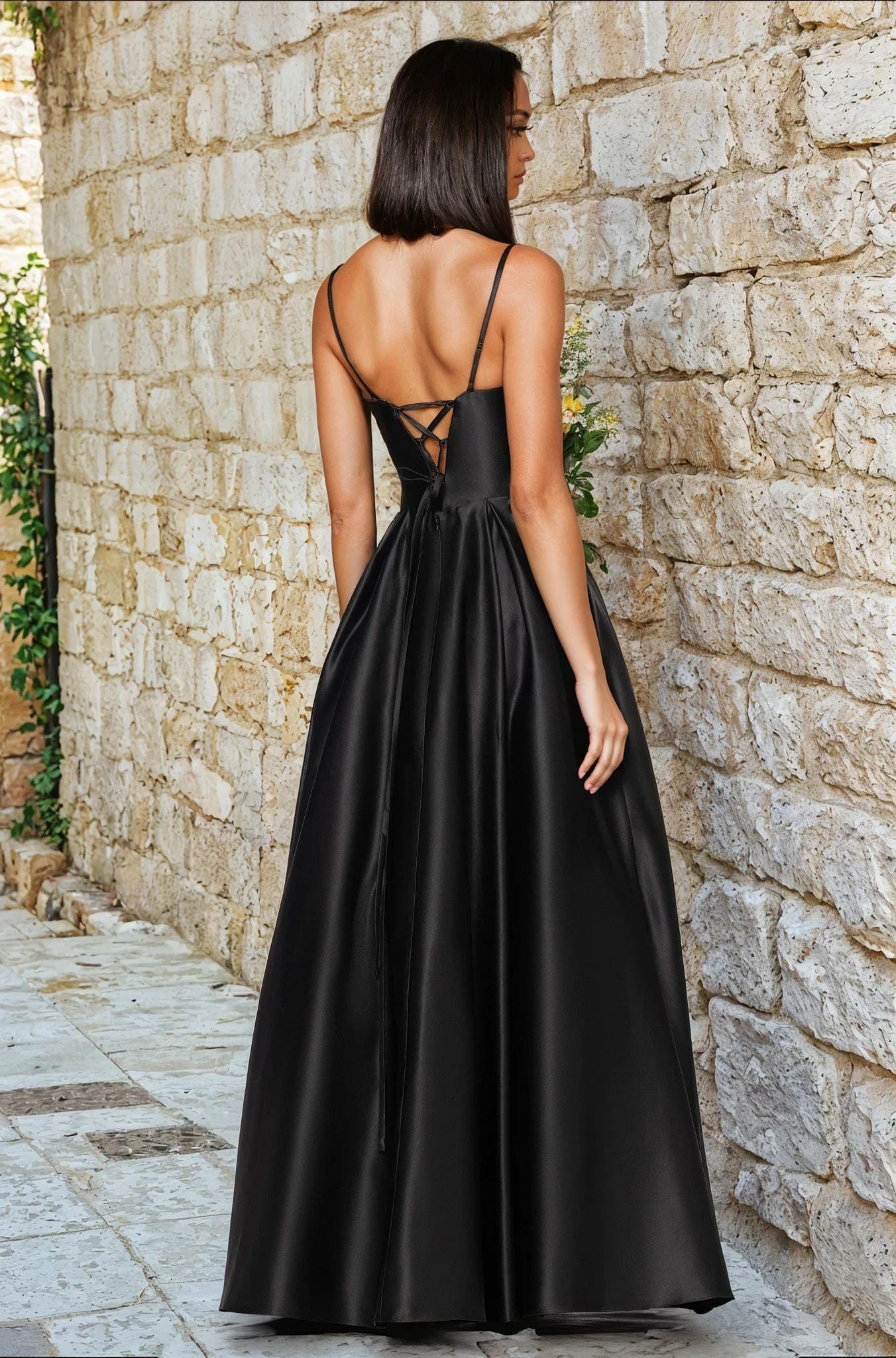 A woman stands on a stone path, facing a stone wall, wearing the Blondie Nites 5453BN long black satin A-line prom dress with thin straps and a lace-up back.