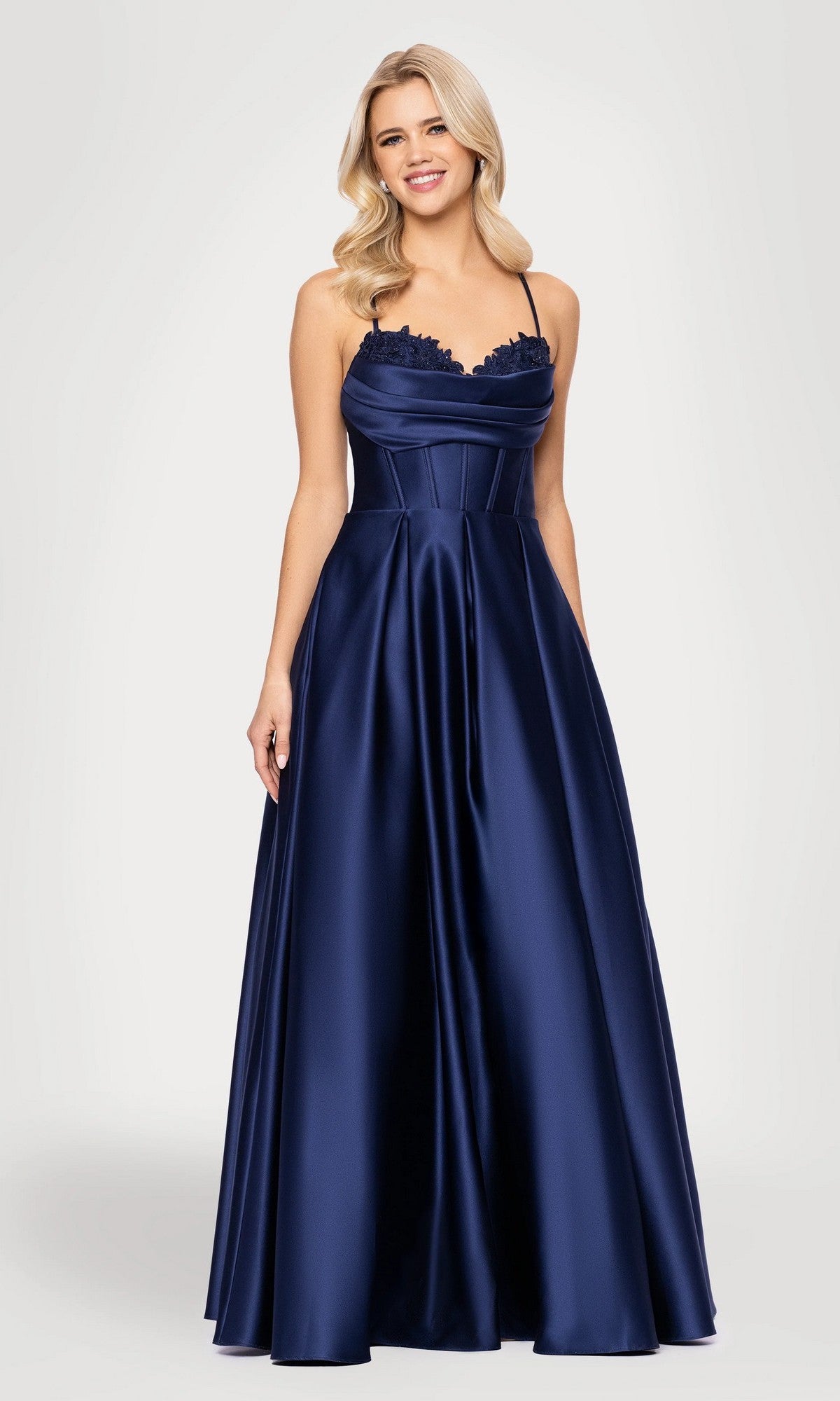 A woman wearing the Long Prom Dress: Blondie Nites 5453BN, a navy blue satin A-line gown with spaghetti straps and a fitted bodice, stands against a plain light background.