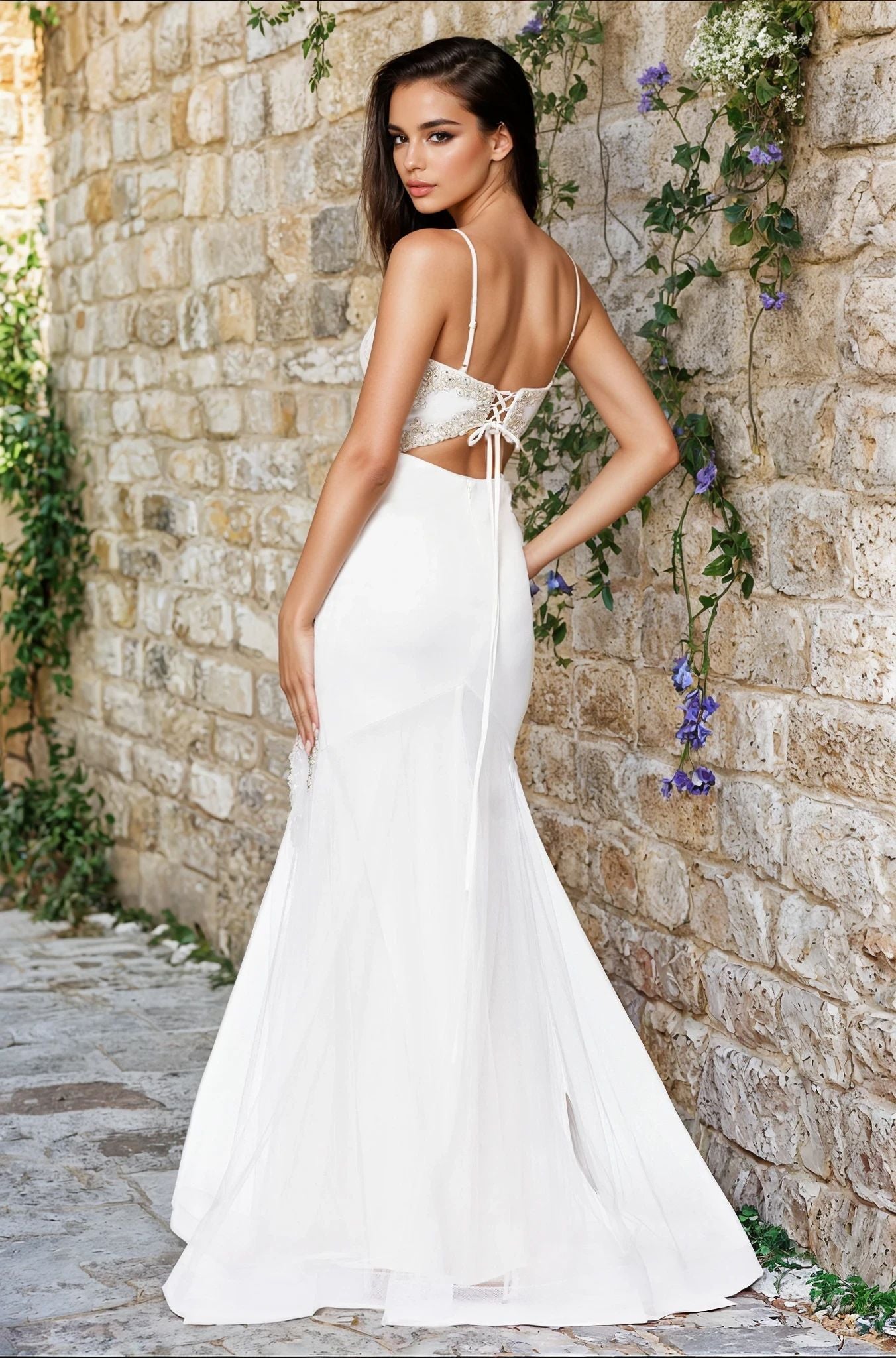 Woman wearing the Blondie Nites 5455BN long prom dress in ivory, featuring a fitted silhouette and lace-up detail, stands on a stone path beside a vine-covered wall, glancing over her shoulder.
