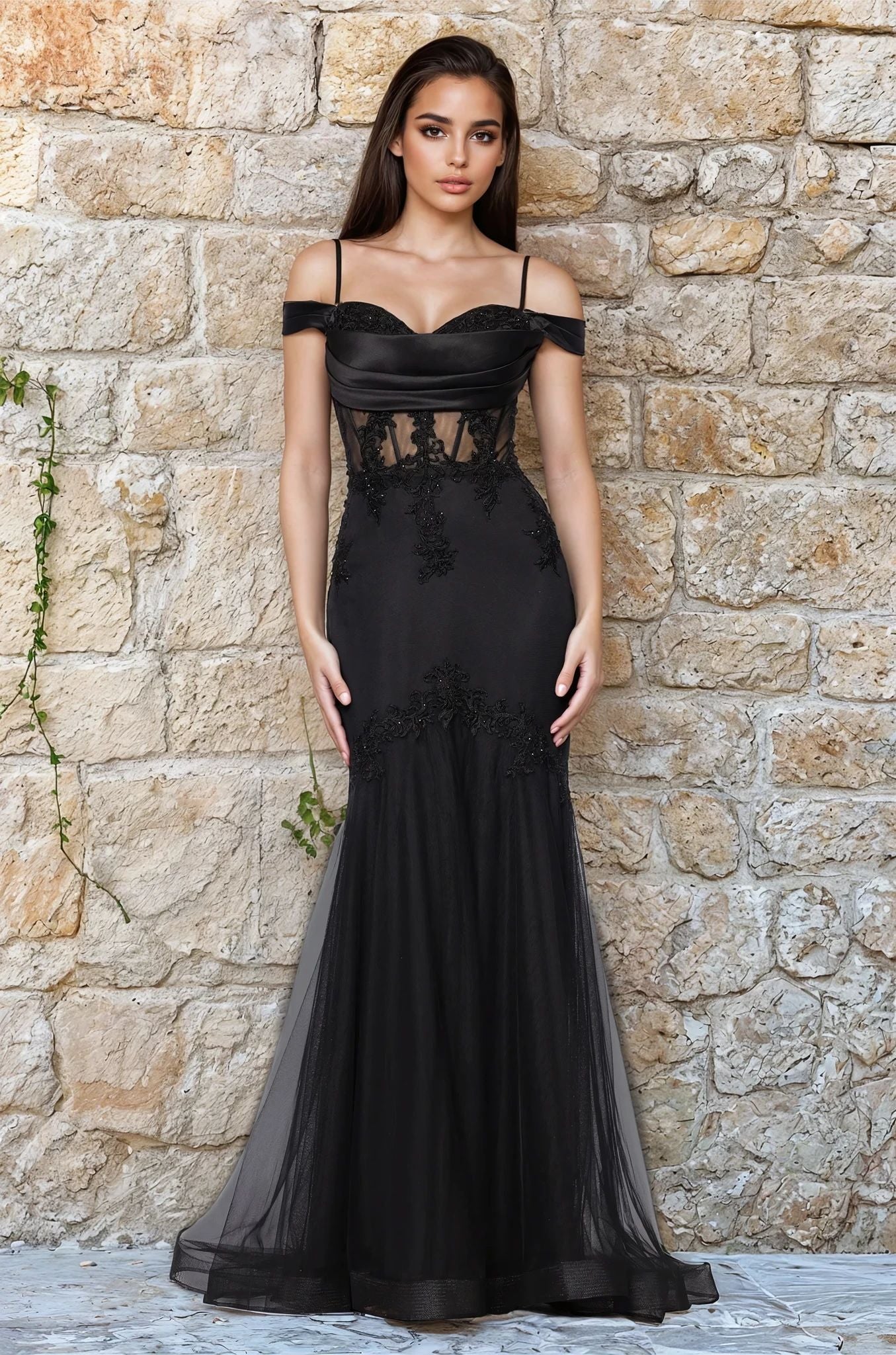 A woman stands against a stone wall, wearing the Black Long Prom Dress: Blondie Nites 5464BN, featuring a striking mermaid silhouette, sweetheart neckline, and sheer panels with delicate lace appliques.