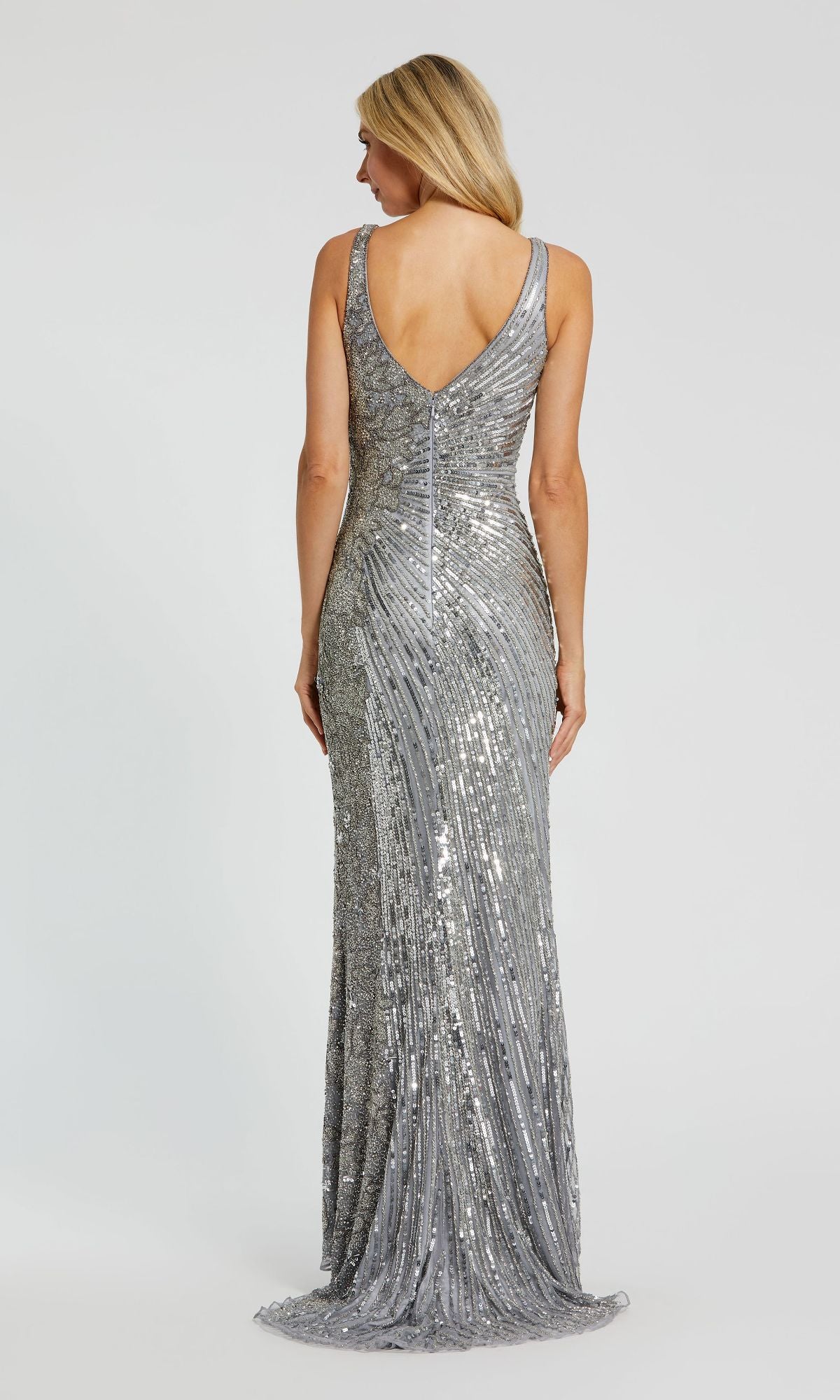 A woman with straight blonde hair stands facing away against a plain light background, wearing the Mac Duggal 5473 Sleeveless Beaded Formal Dress featuring shimmering beads and a V-shaped back.