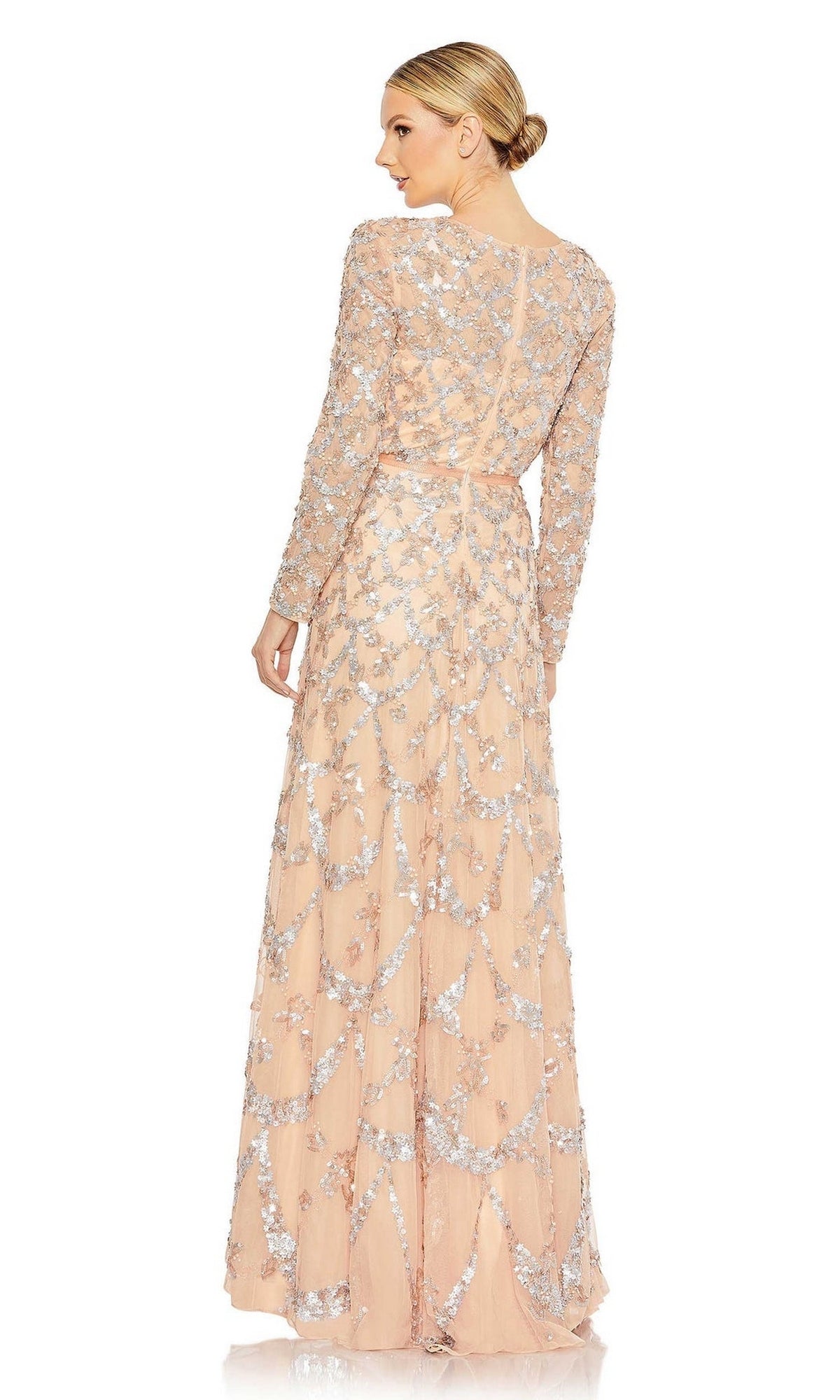A woman faces away in the Mac Duggal 5496 long sleeve sequin formal gown. The floor-length blush dress features silver sequin floral patterns, offering an elegant and graceful mother of the bride look.