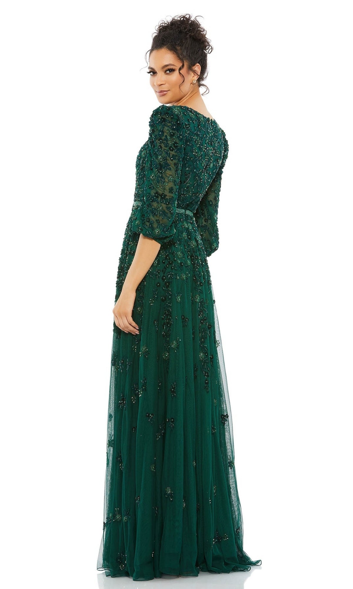 A woman stands looking over her shoulder in the Mac Duggal 5497, a floor-length emerald green formal dress with long sleeves, beading, and floral appliqué.