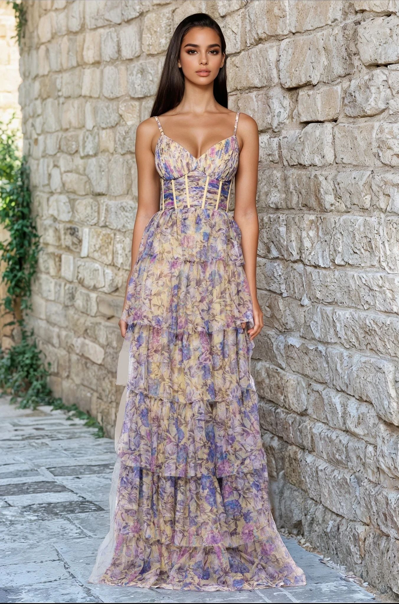 A woman stands on a stone walkway against a stone wall, wearing the Blondie Nites 5500BN Floral-Print Long Prom Dress featuring tiered layers and a corset-style bodice.
