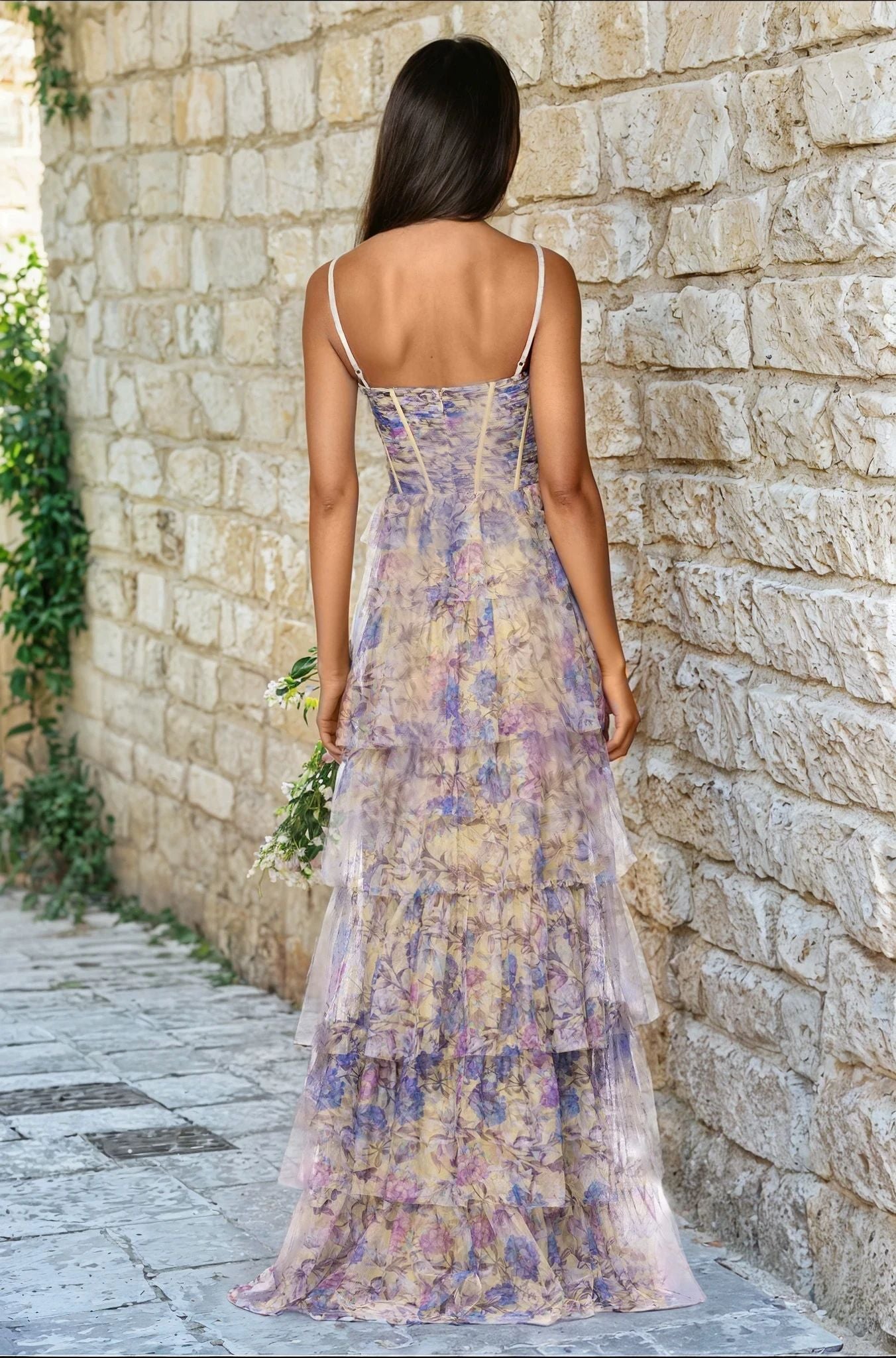 A woman with long brown hair wears the Blondie Nites 5500BN Floral-Print Long Prom Dress with thin straps, standing on a stone path by a wall, facing away from the camera.