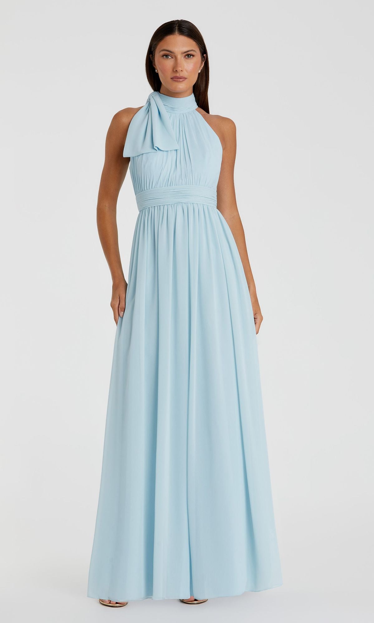 A woman stands against a plain white background wearing the sleeveless, floor-length light blue Mac Duggal 55035 high-neck long formal dress with bow detail.