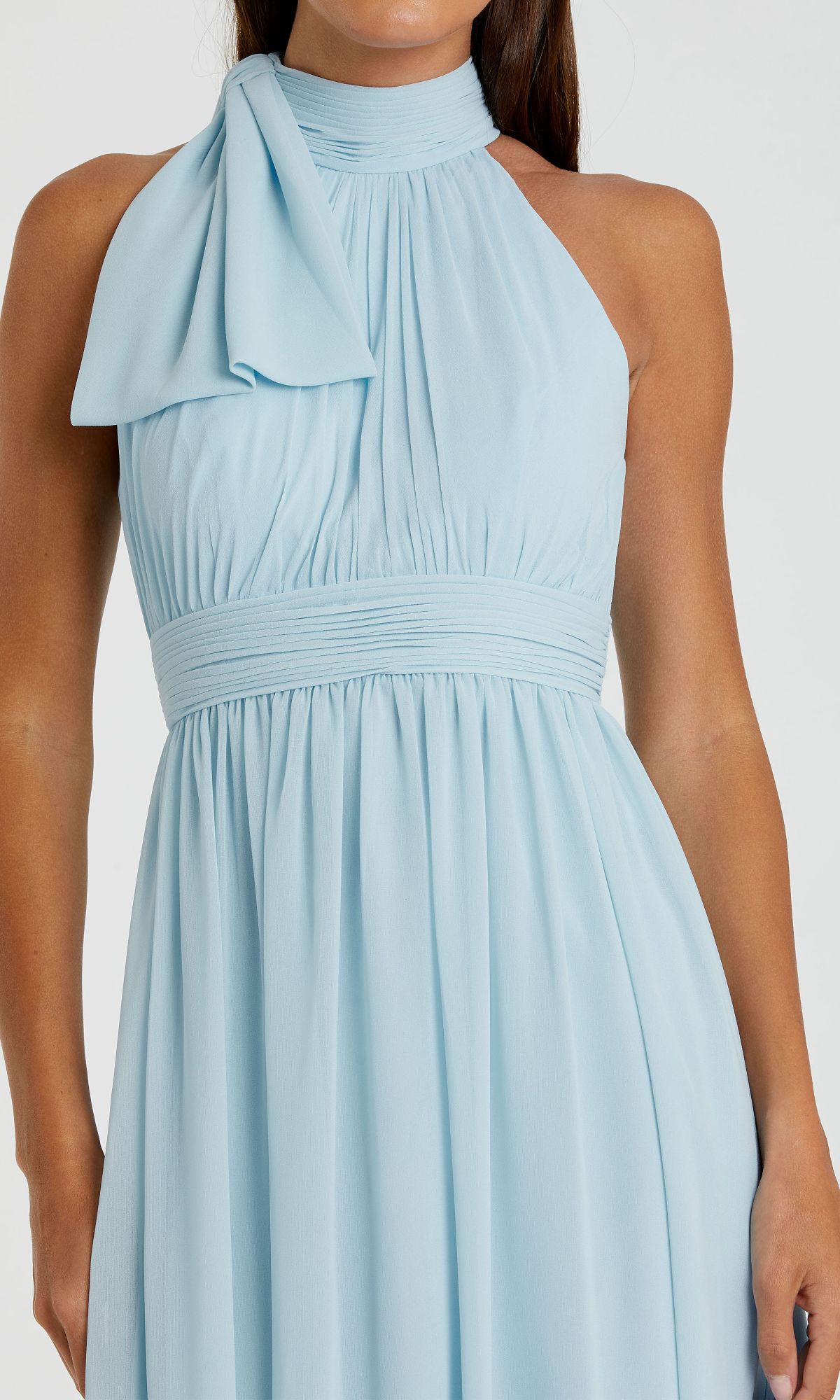 A woman in the Mac Duggal 55035 High-Neck Long Formal Dress, light blue and sleeveless, with a halter neck, pleated bodice, and a one-shoulder bow detail.