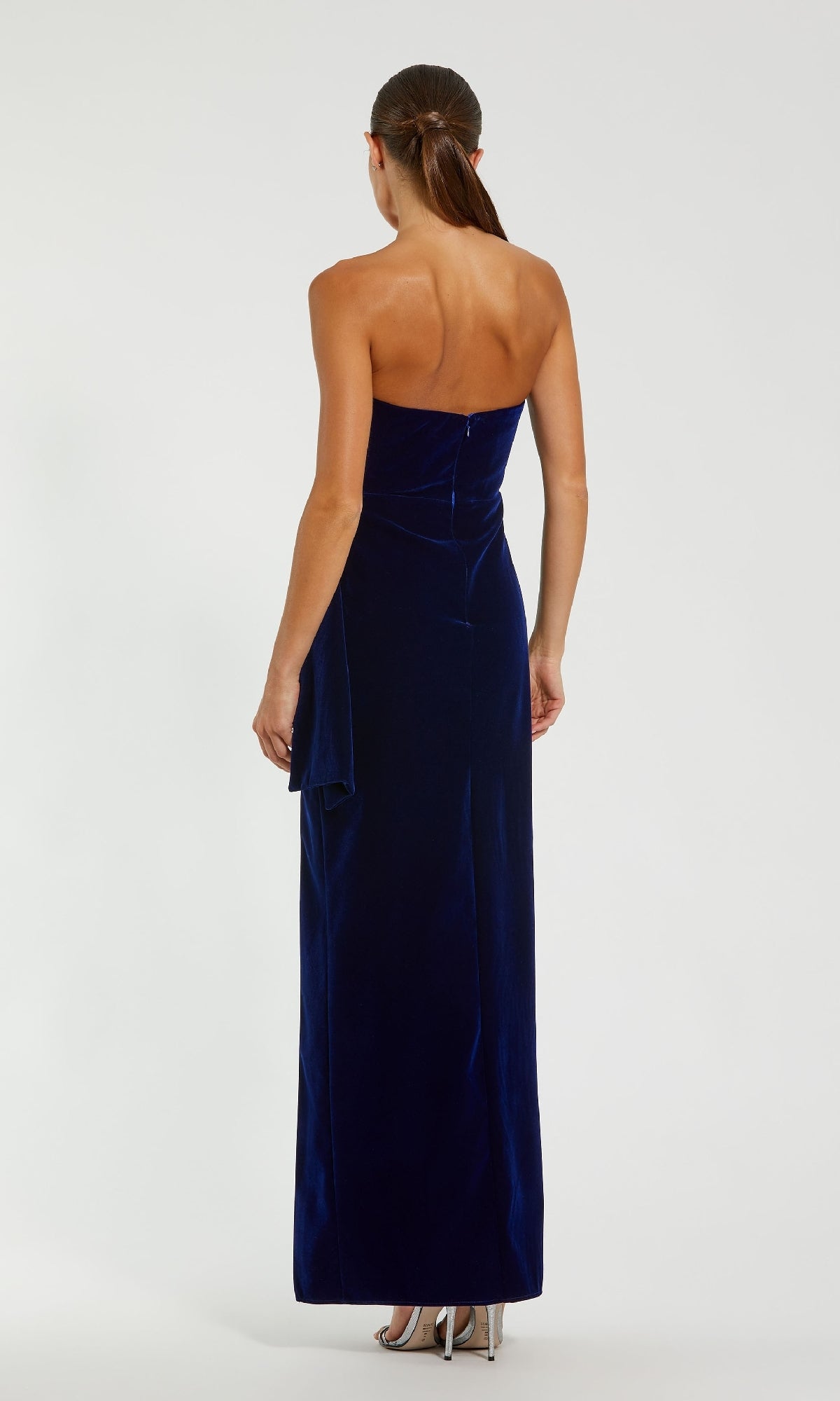 A woman stands facing away, wearing the Mac Duggal 55080 Strapless Velvet Formal Gown in dark blue with silver high heels against a plain light background.