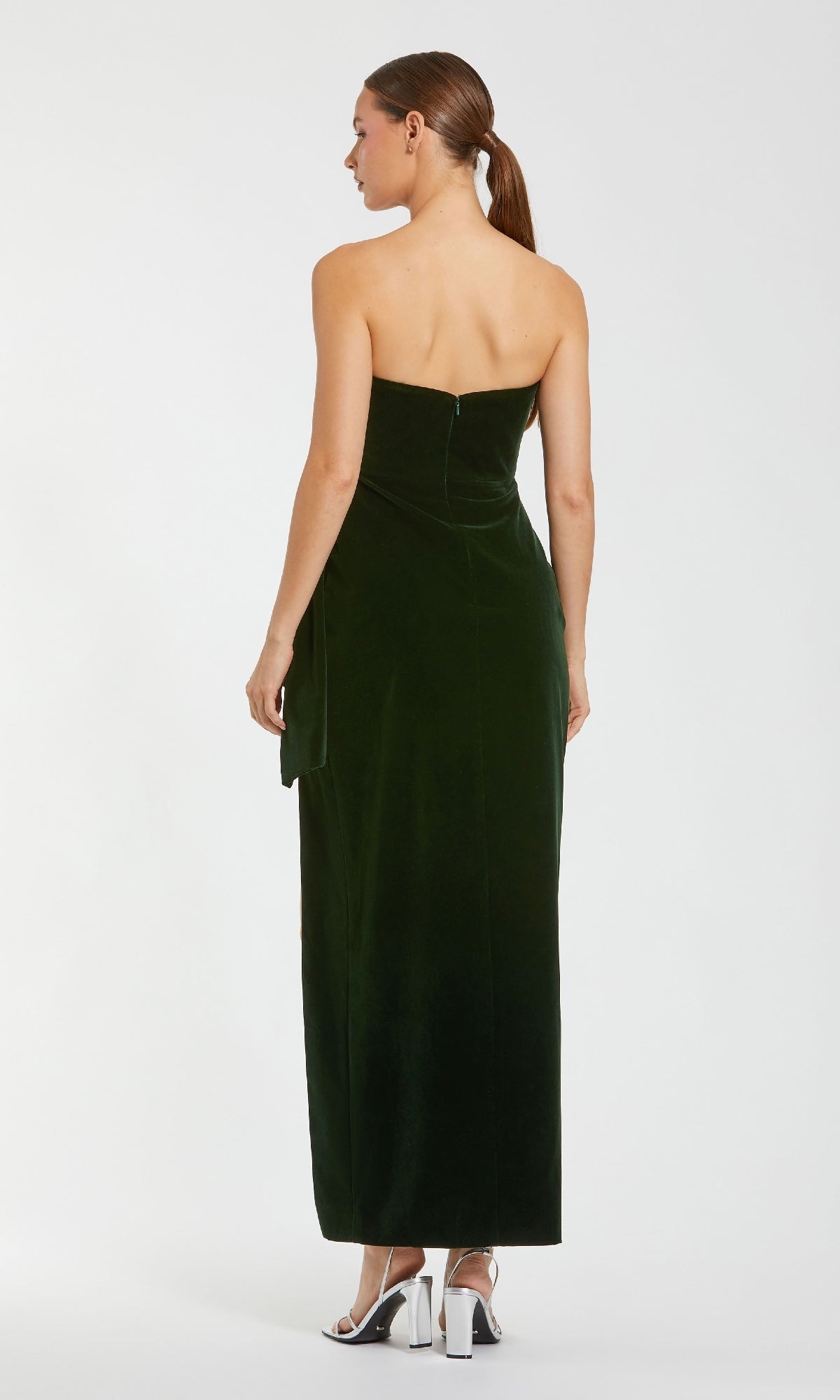 A woman, facing away with a low ponytail and silver heels, wears the Mac Duggal 55080 Strapless Velvet Formal Gown in dark green, featuring side detailing.