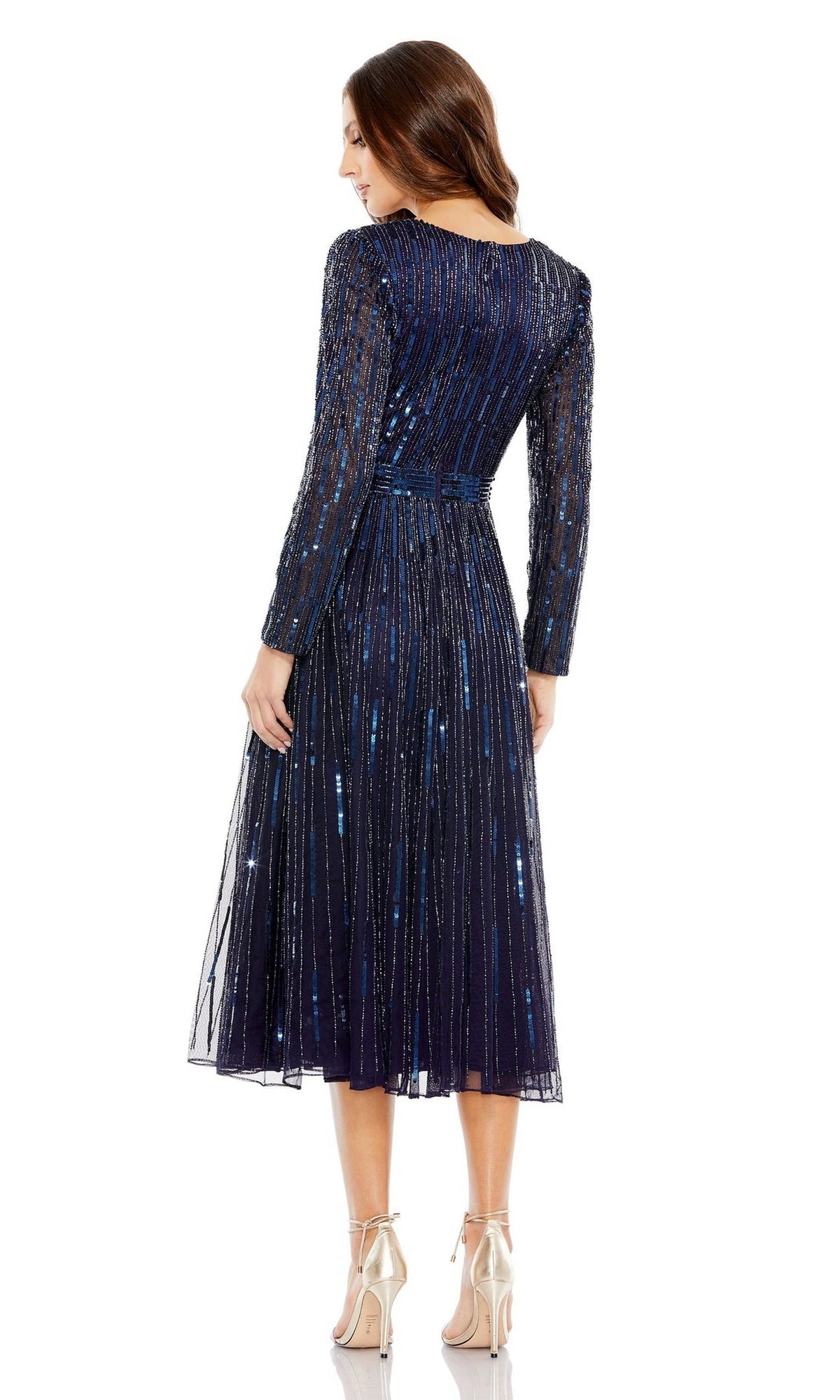 A woman stands facing away in the Short Semi-Formal Dress 5528 by Mac Duggal, featuring long sleeves, navy blue sequins, an A-line tea length silhouette with a pleated skirt, paired with silver high heels.