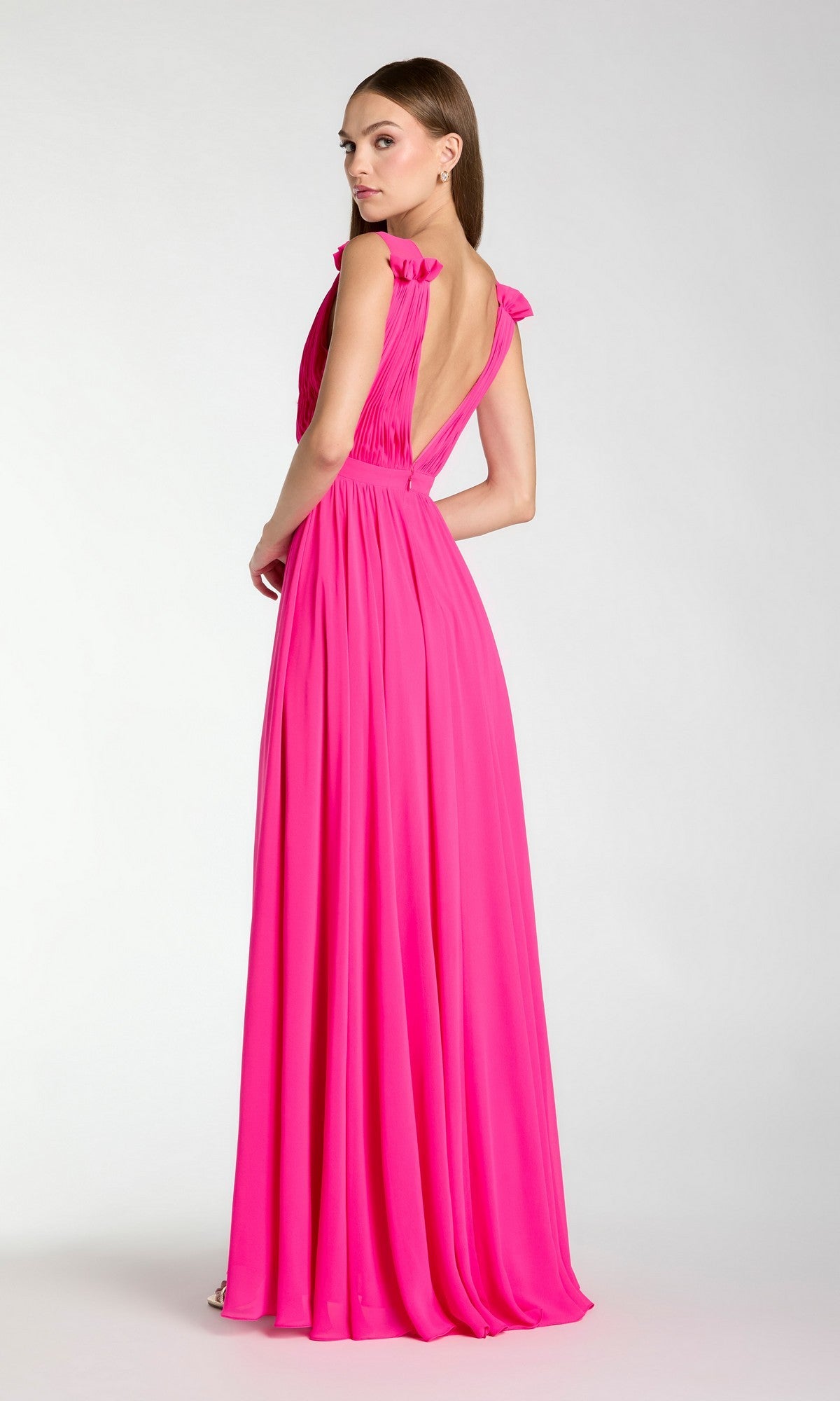 A woman stands against a plain background, looking over her shoulder in the Classic Long Formal Dress: Mac Duggal 55321—a sleeveless, floor-length bright pink evening gown with an open back.