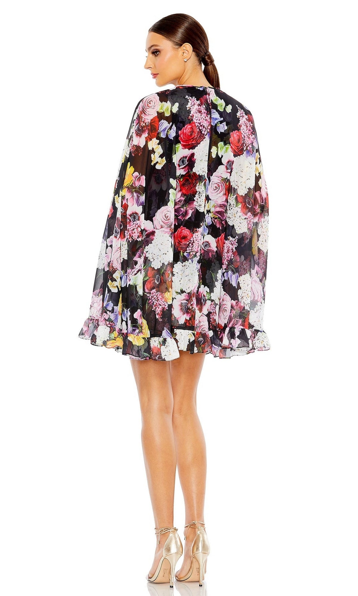 A woman stands facing away, wearing the Mac Duggal 55404 Short Print Cape Hoco Dress with a ruffled hem and high-heeled sandals.
