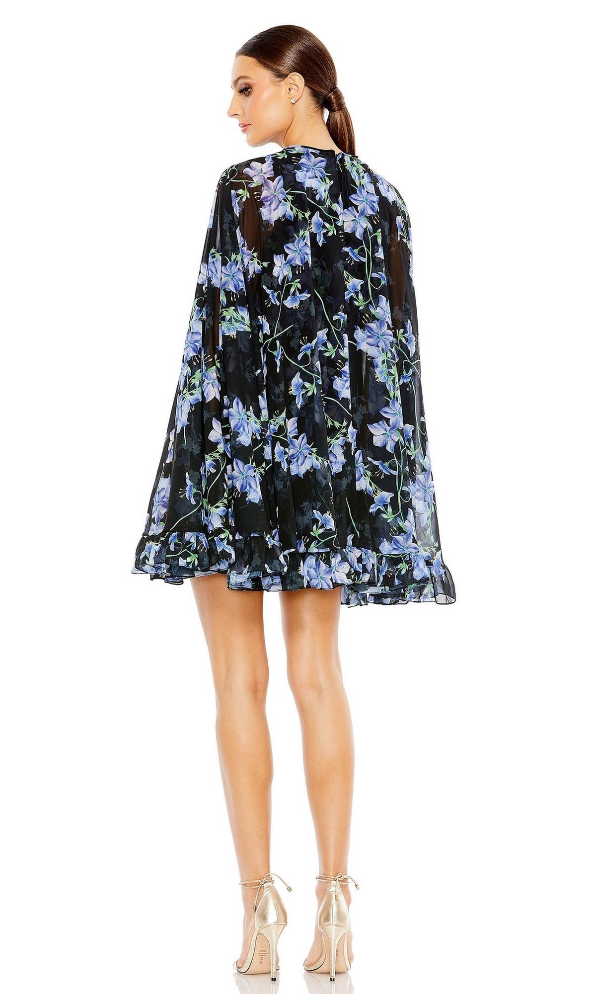 A woman faces away in a Mac Duggal 55404 Short Print Cape Hoco Dress with a black floral print, blue flowers, flowy silhouette, ruffled hem, and beige high-heeled sandals.