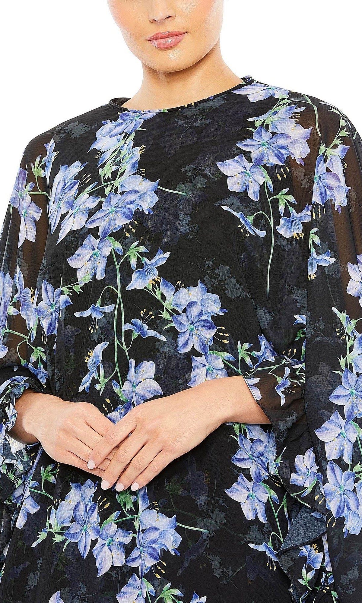 A woman models the Mac Duggal 55404 Short Print Cape Hoco Dress, featuring a black base with blue and purple floral patterns, shown from shoulders to mid-torso with hands clasped in front.