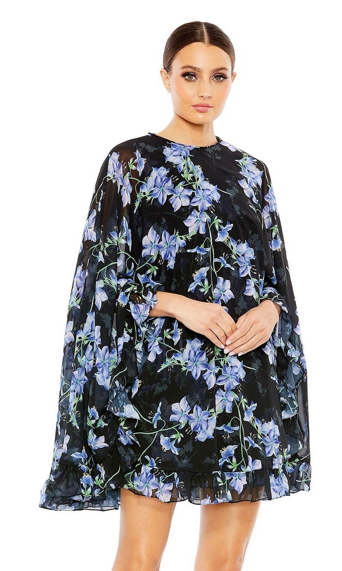 A woman wears the Mac Duggal 55404 Short Print Cape Hoco Dress—black with blue and green floral patterns and loose-fitting long sleeves—standing against a white background.