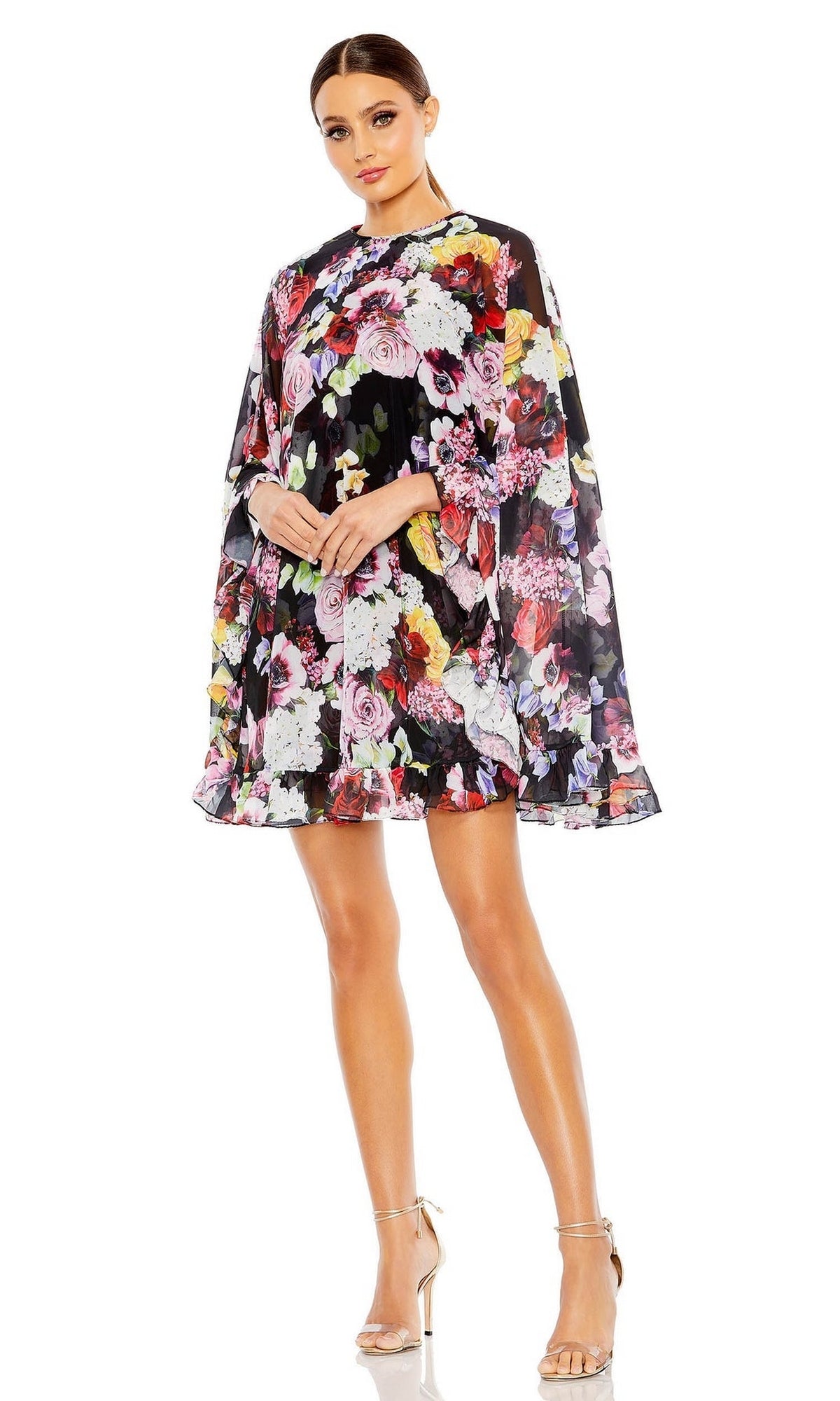 A woman wears the Mac Duggal 55404 Short Print Cape Hoco Dress with floral print and long, cape-like sleeves, paired with silver high-heeled sandals.